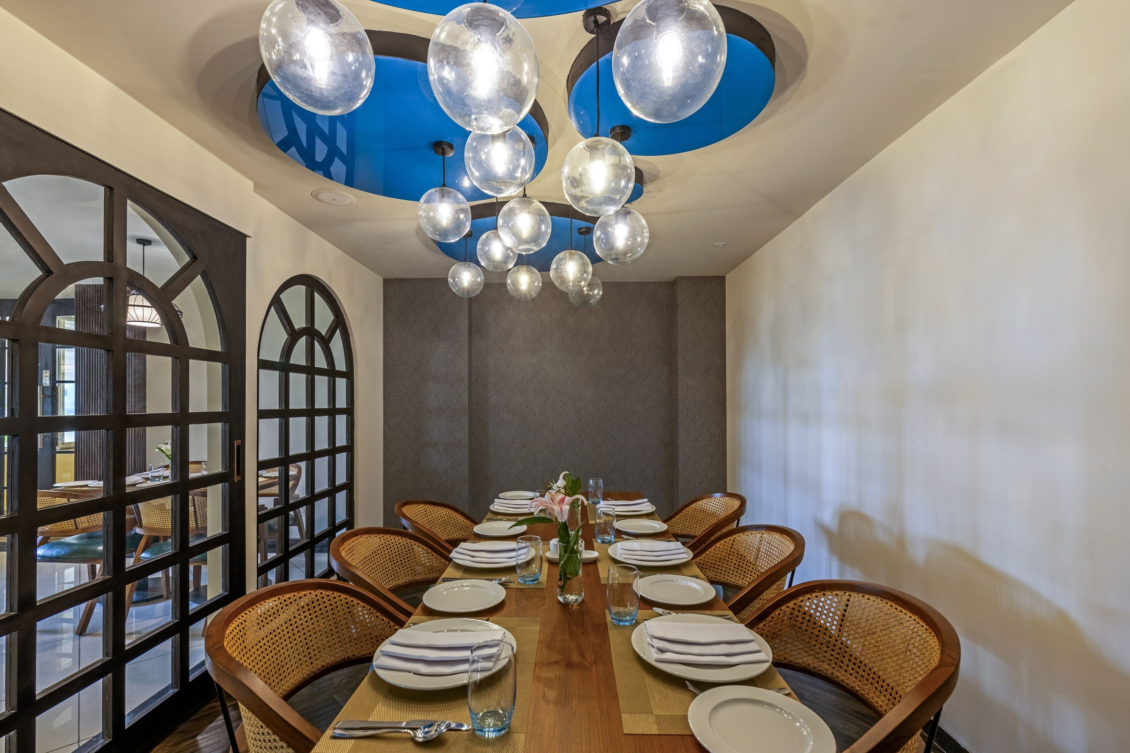 Zona pranzo con un lungo tavolo in legno apparecchiato per un pasto, circondata da sedie in vimini e un soffitto con lampade a sfera.