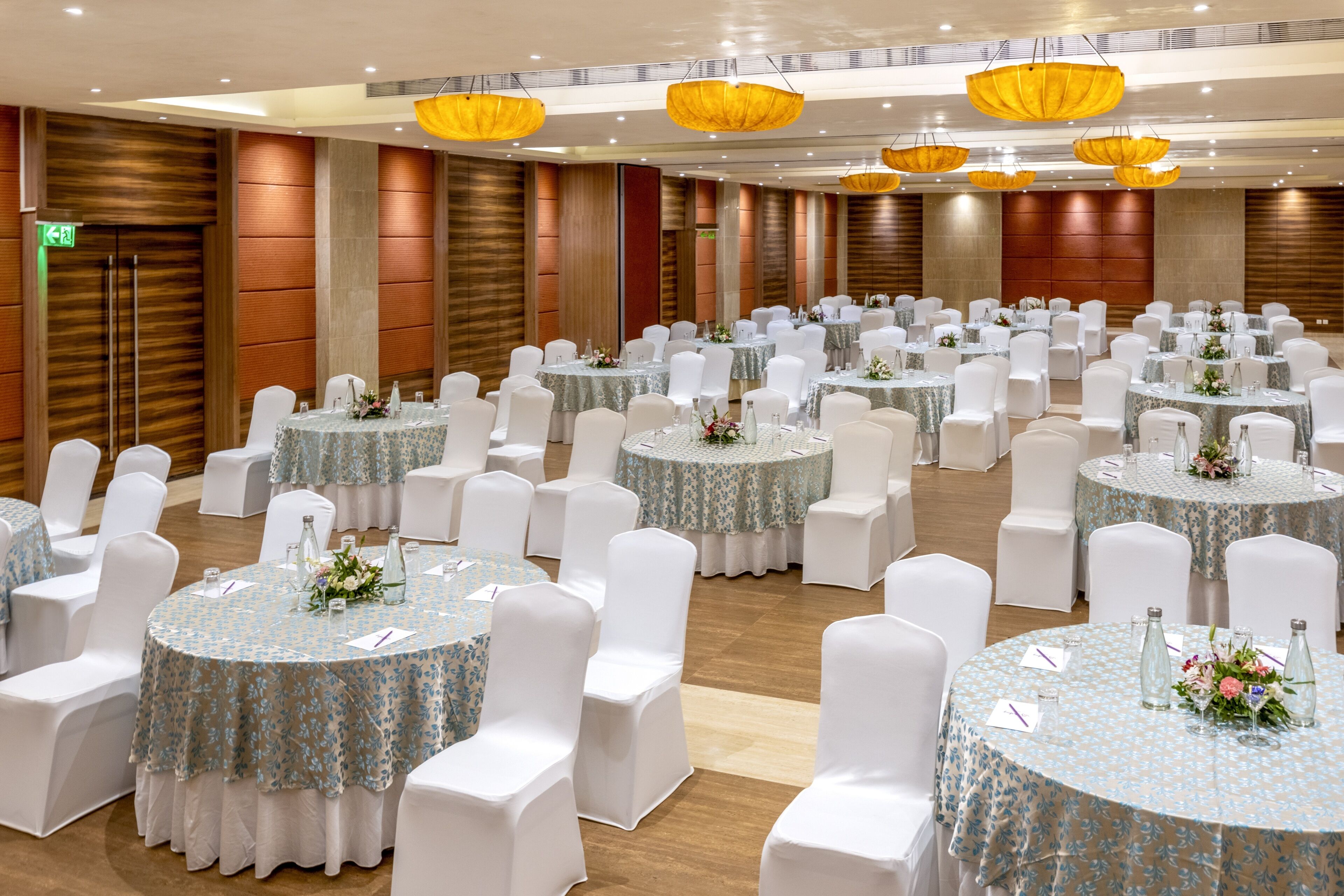 Uma sala de banquetes com mesas redondas, cadeiras brancas e arranjos florais.