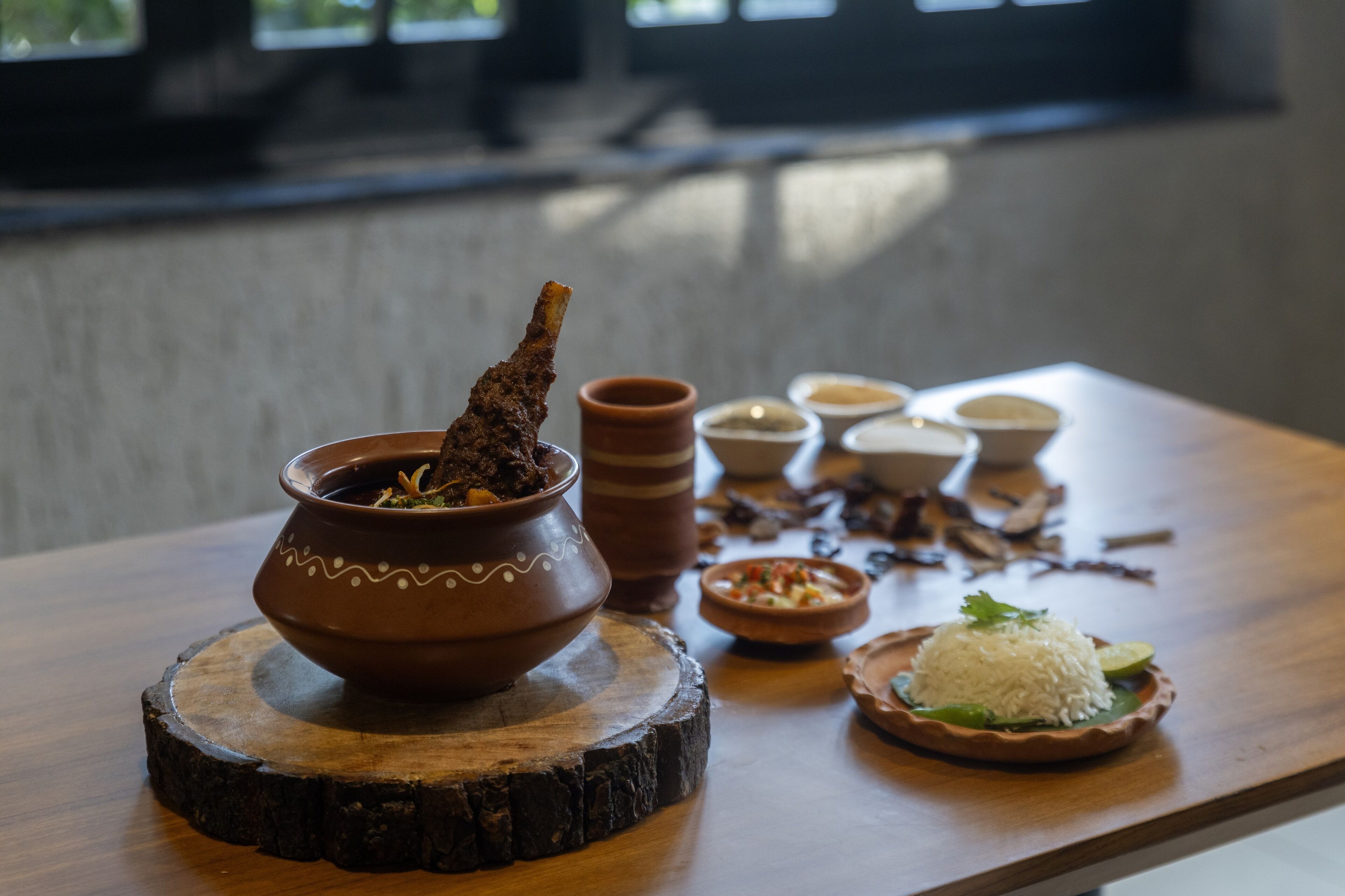 Una mesa de madera con una olla de barro que contiene comida, un plato de arroz con decoración, y varios recipientes pequeños con salsas.
