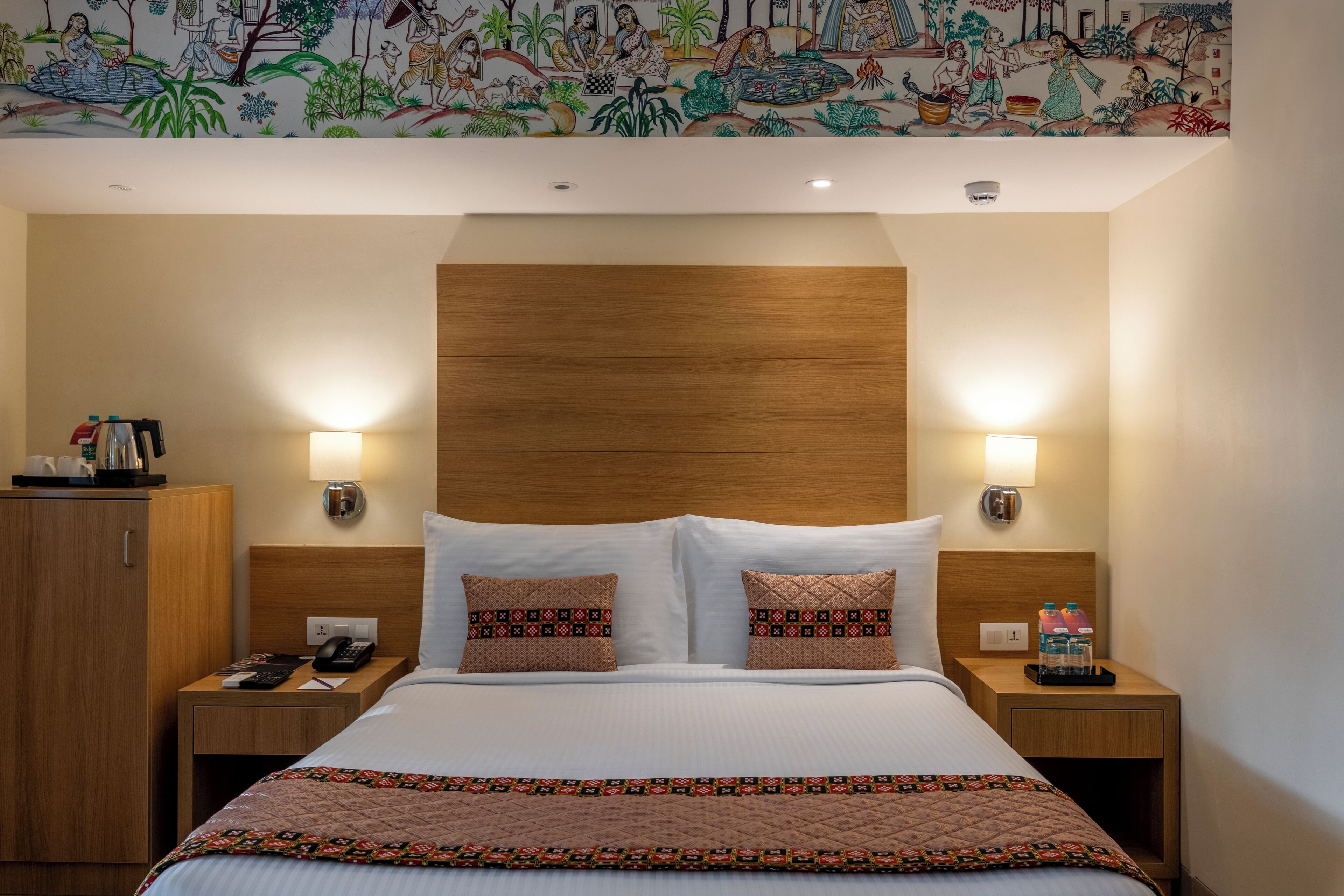 A hotel room with a wooden headboard, two bedside tables with lamps, a bed with patterned pillows, and a colorful mural on the wall.