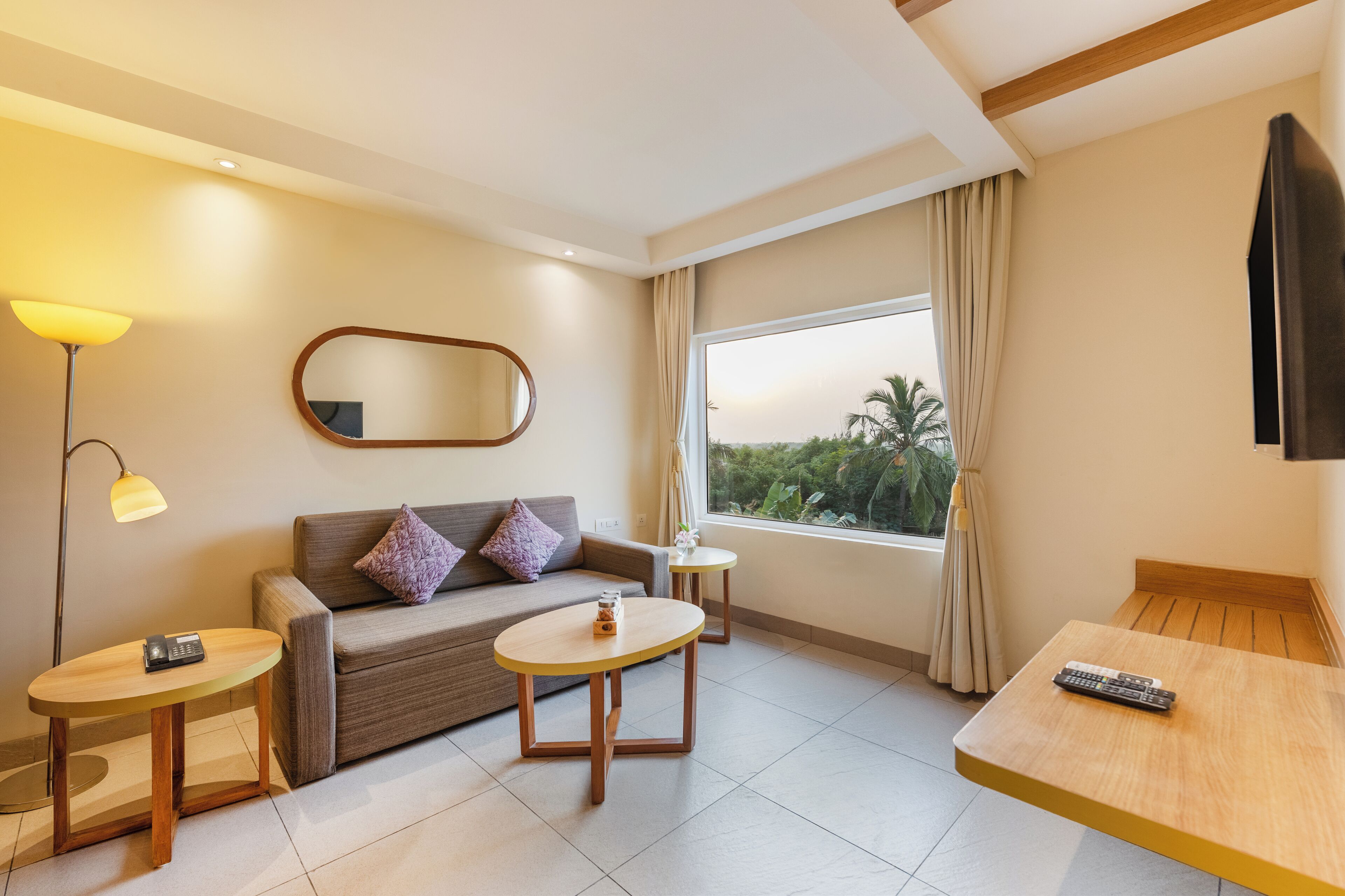 A modern hotel room with a sofa, two small tables, a large window with a view of greenery, and a flat-screen TV mounted on the wall.