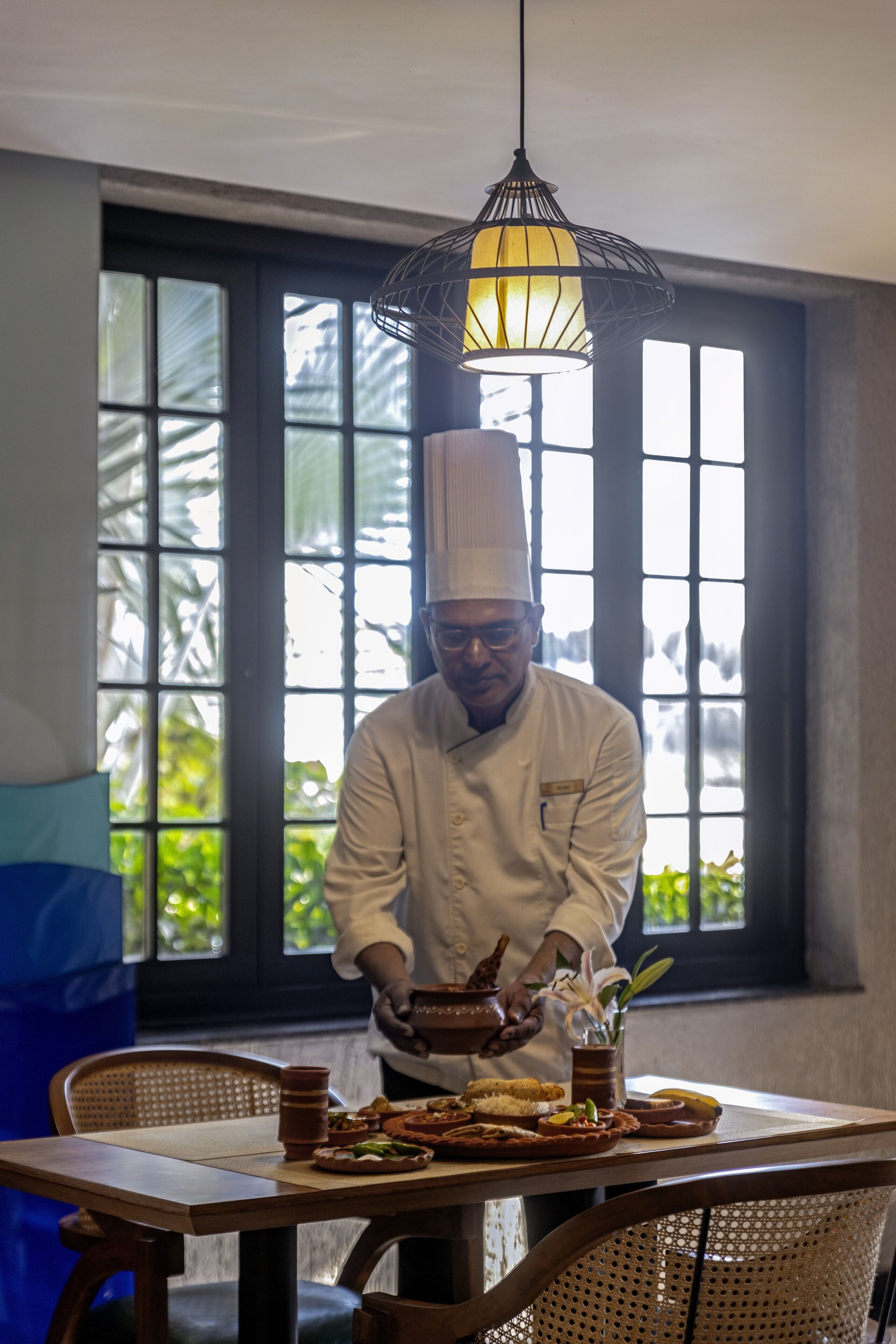 A chef in a white uniform preparing food at a table with a traditional clay pot and a decorative vase.