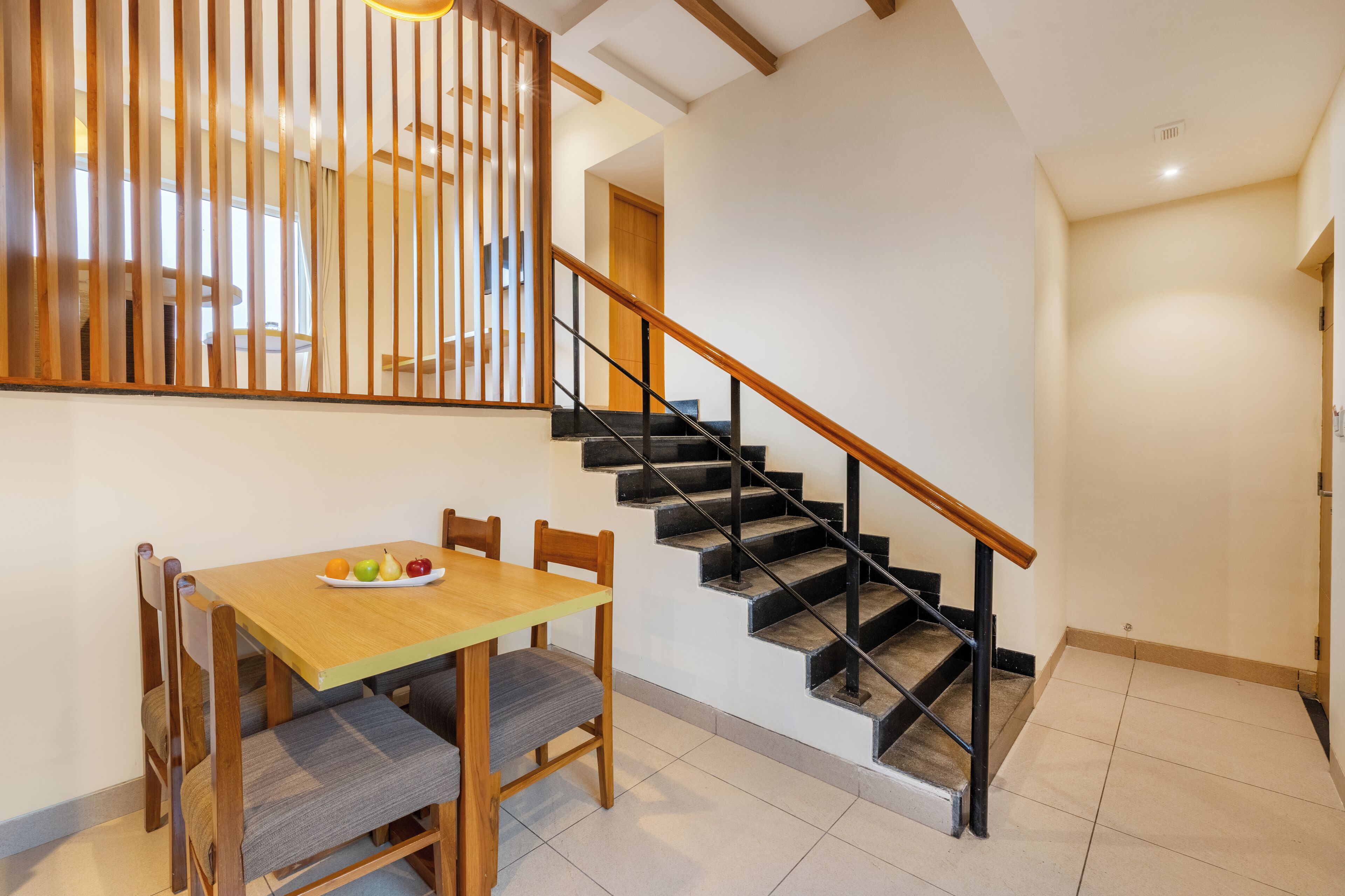 A dining area with a wooden table and chairs, a fruit bowl, and a staircase with black railings.