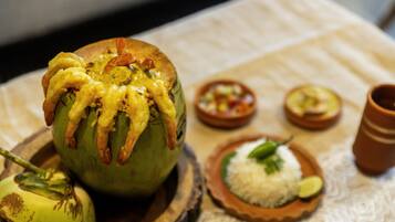Una mesa de comedor con una fruta verde grande, posiblemente un guayaba, coronada con camarones y salsa, acompañada de un plato de arroz blanco y un tazón pequeño de vegetales mixtos.