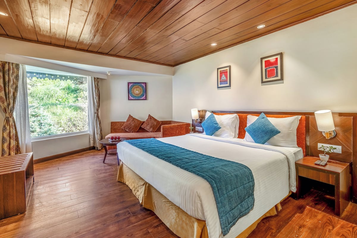 A spacious bedroom with a large bed, a wooden headboard, a bedside table with a lamp, and a view of greenery through a window.