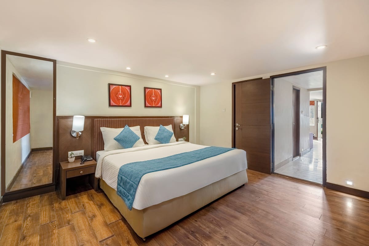 A hotel room with a large bed, wooden flooring, a nightstand with a lamp, and two framed pictures on the wall.