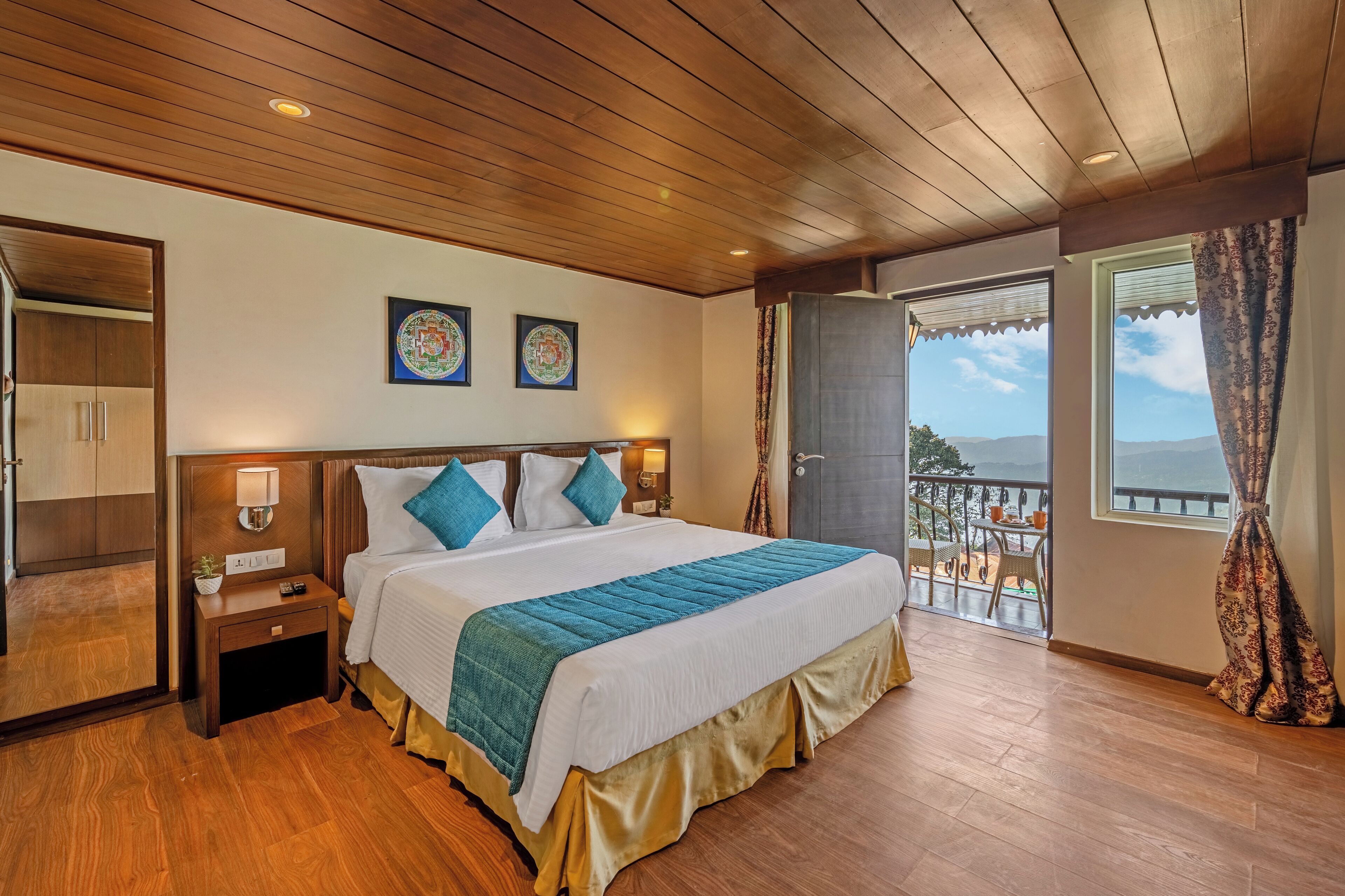 Habitación de hotel con techo de madera, una cama grande con colcha azul y blanca, una mesita de noche con lámpara y vistas al exterior a través de una puerta de balcón.