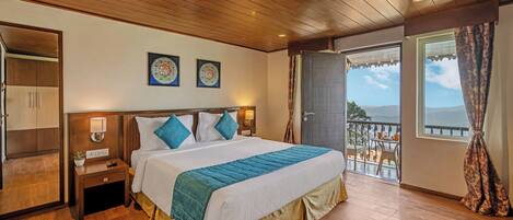 Habitación de hotel con techo de madera, una cama grande con colcha azul y blanca, una mesita de noche con lámpara y vistas al exterior a través de una puerta de balcón.