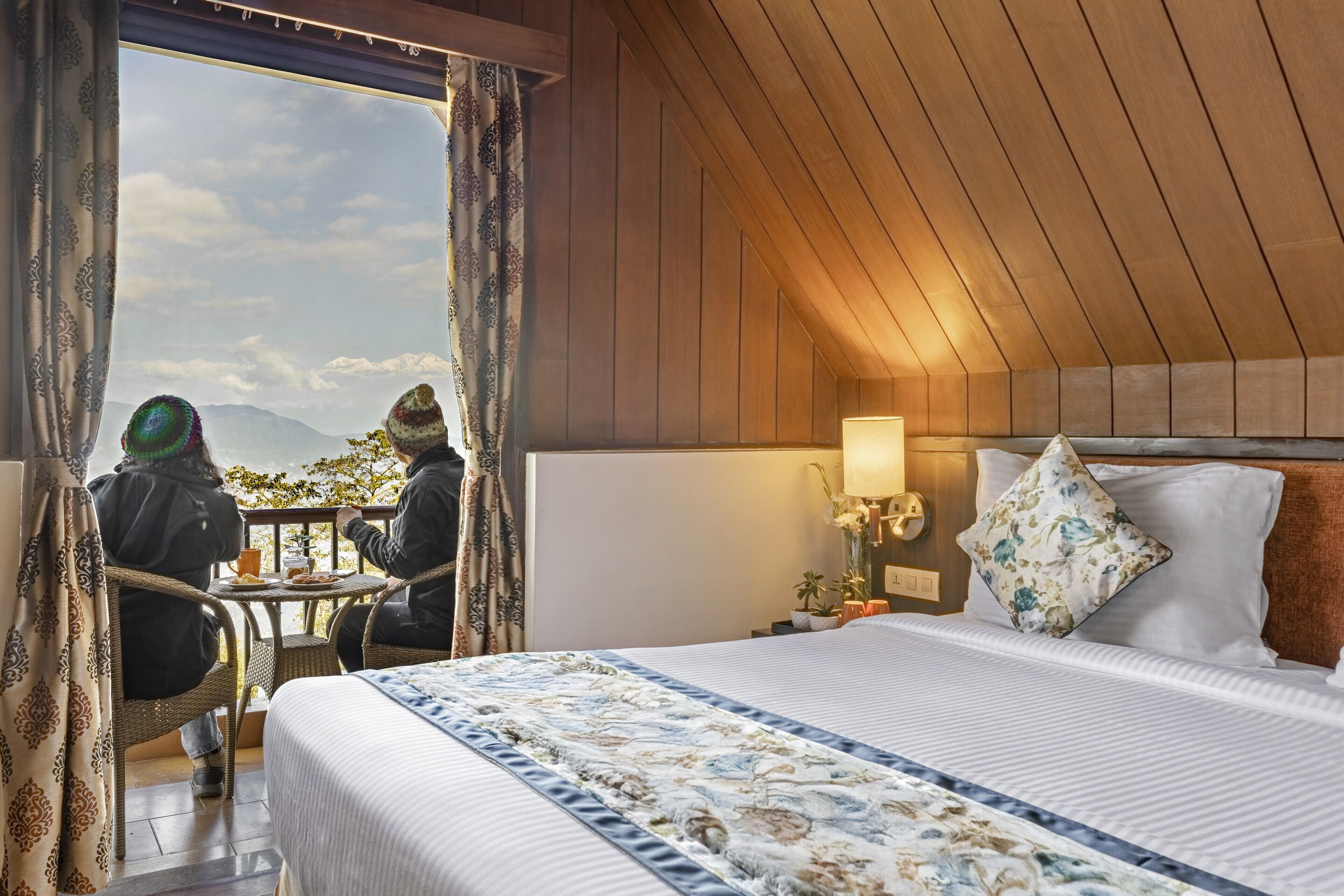 Een hotelkamer met een bed, een nachtkastje, een lamp en uitzicht op bergen door een raam.