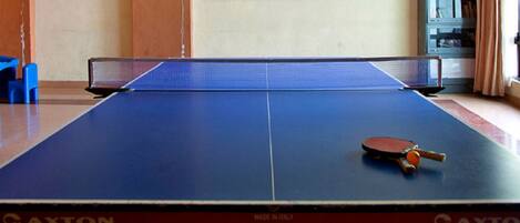 Een tafeltennistafel met een racket in het midden, een blauwe stoel links en een boekenkast op de achtergrond.