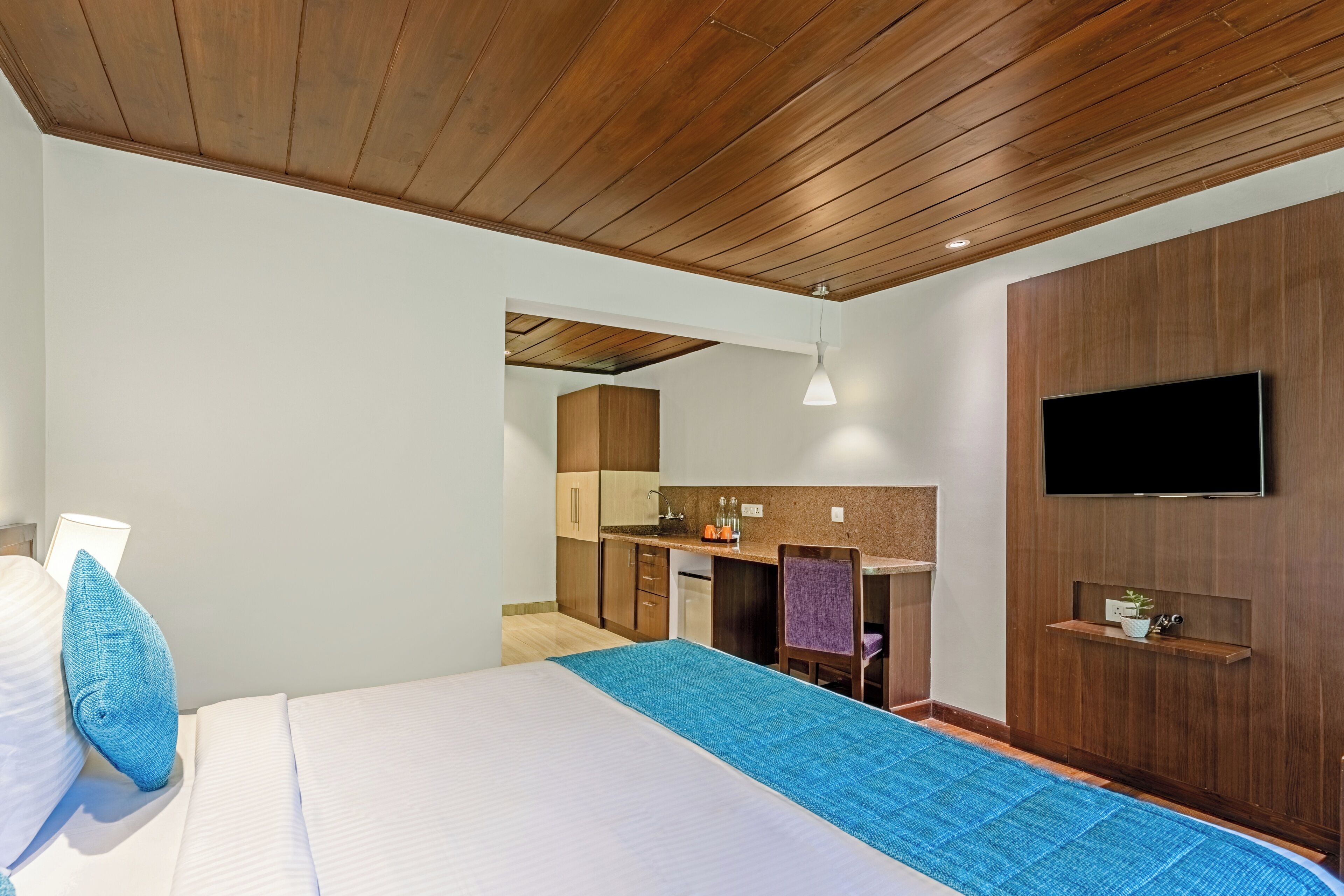 A modern hotel room with a wooden ceiling, a large bed with a blue runner, a TV mounted on the wall, and a kitchenette in the background.