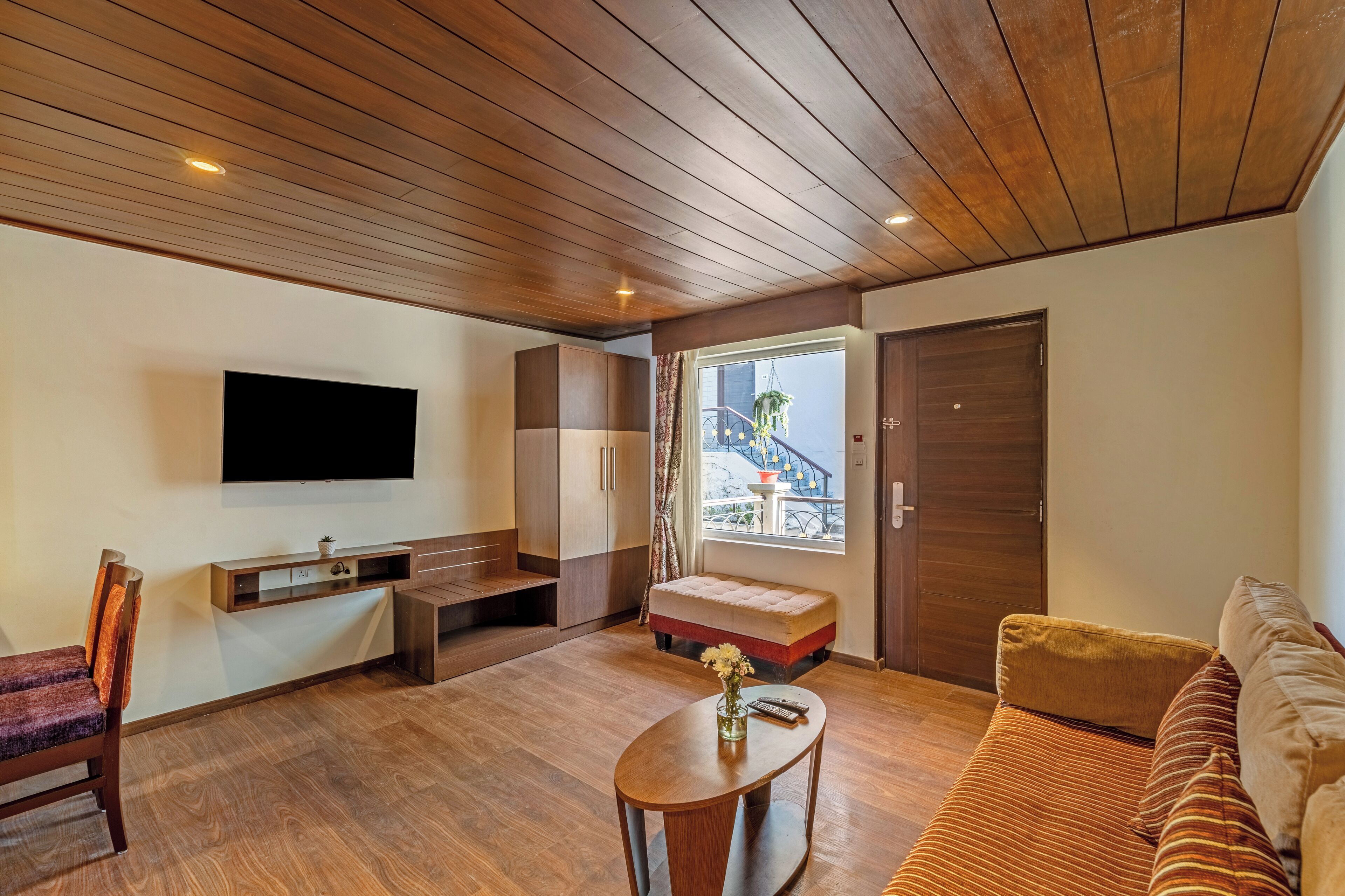 A modern living room with a wooden ceiling, a flat-screen TV mounted on the wall, a sofa, a coffee table, and a window with a view of a balcony.
