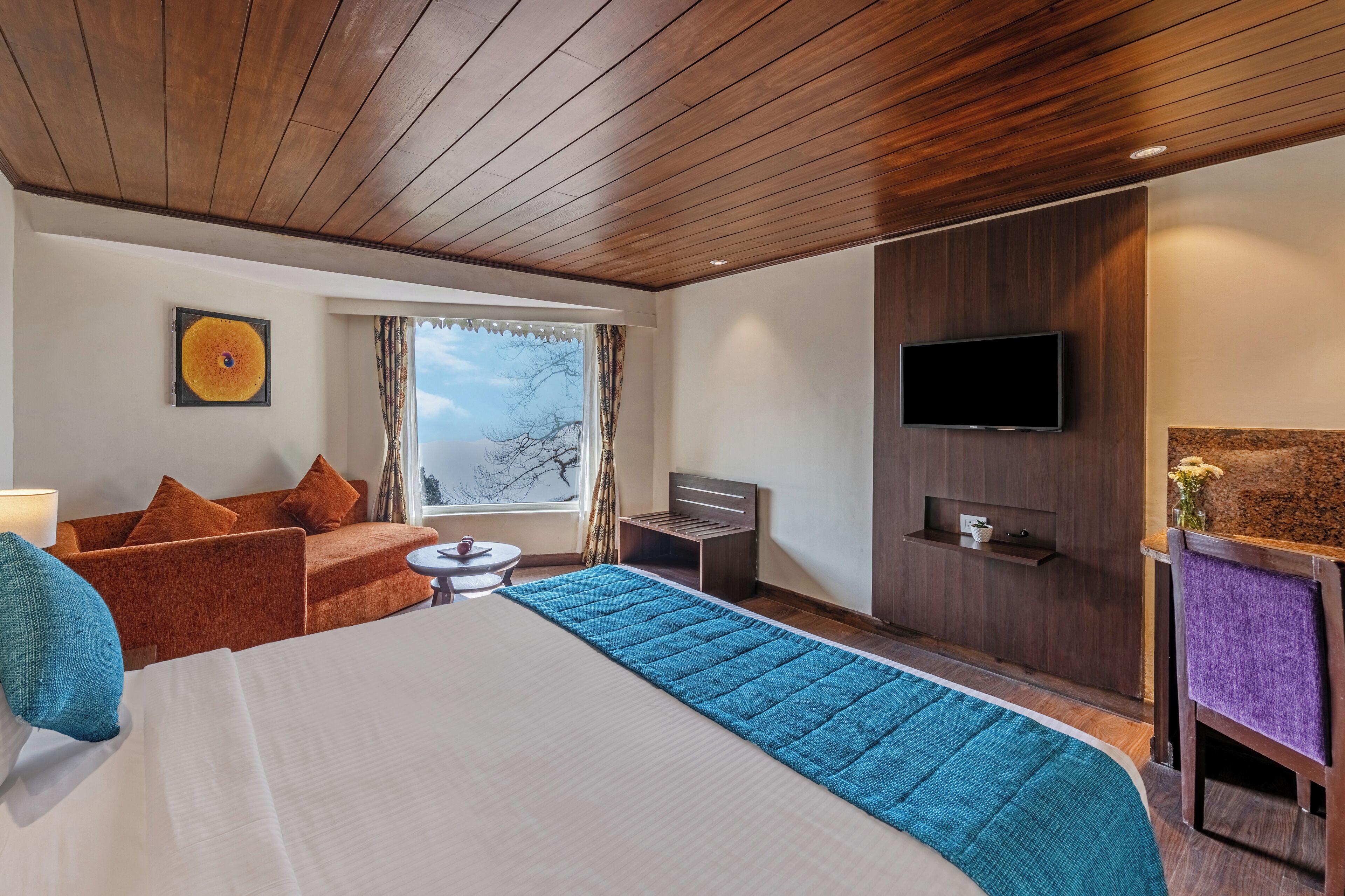 A hotel room with a wooden ceiling, a bed with a blue bedspread, a sofa with orange cushions, a small table, a TV mounted on the wall, and a window with a view of trees.