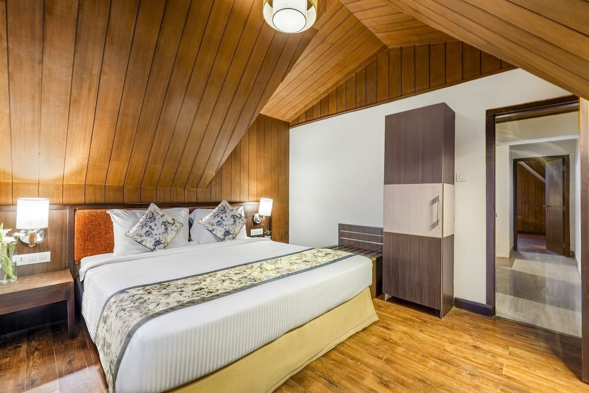 A bedroom with a wooden ceiling, a large bed with a floral bedspread, a nightstand with a lamp, and a wardrobe.