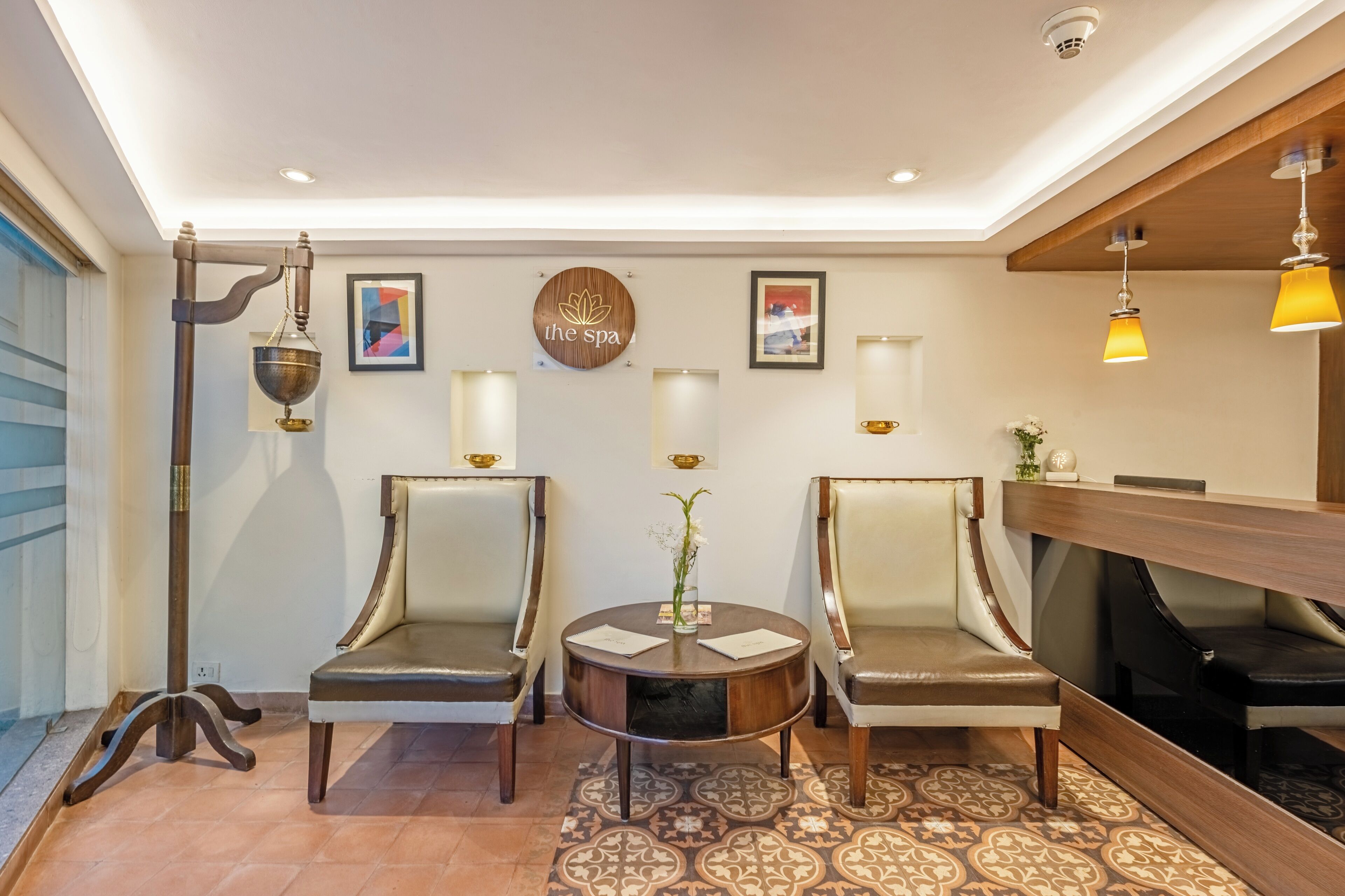 Un área de recepción con dos sillones de cuero, una mesa redonda con un florero, luces en la pared y un mostrador de recepción de madera.