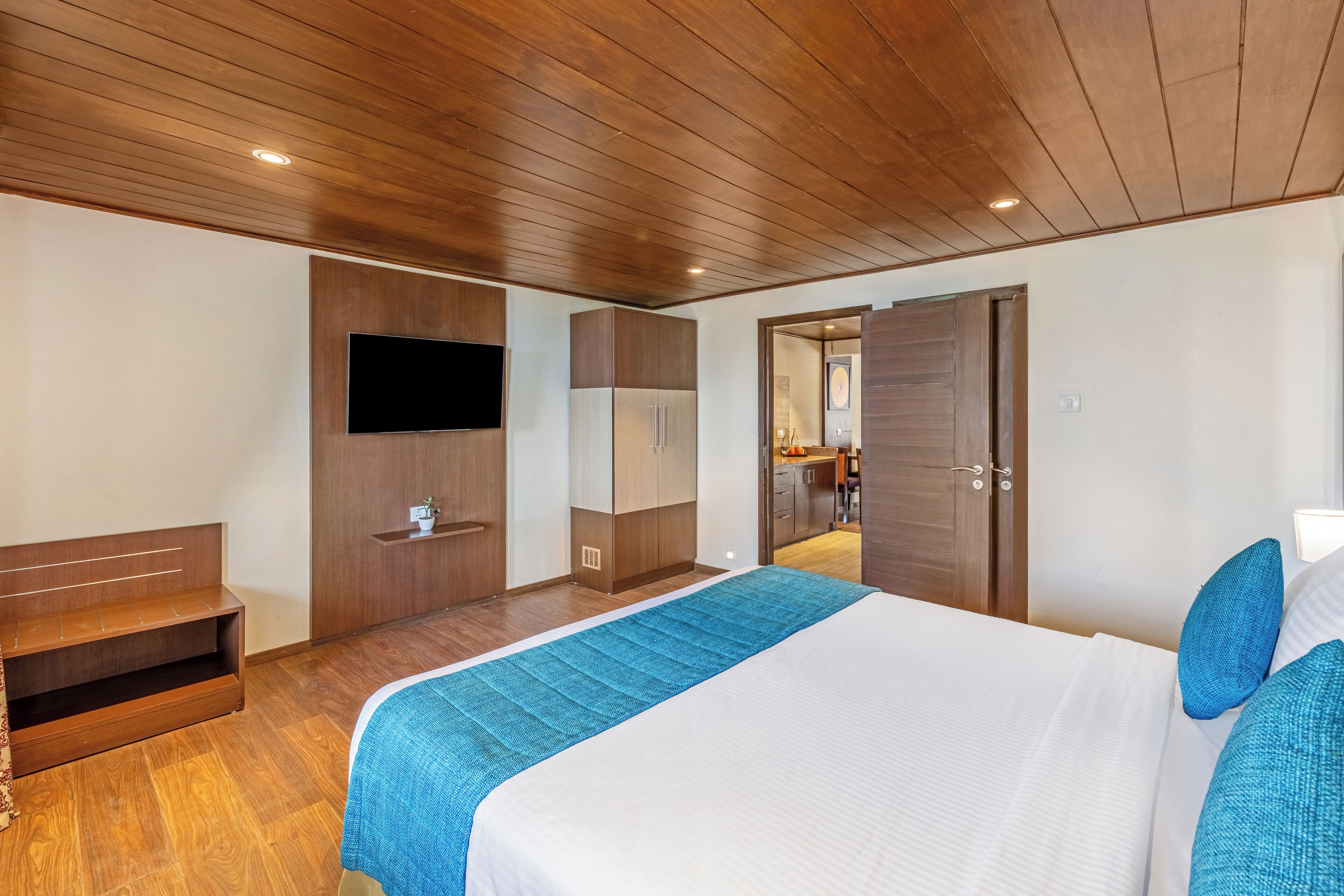 Una habitación de hotel moderna con techo de madera, una cama grande con ropa de cama azul, un televisor de pantalla plana en la pared y un armario.
