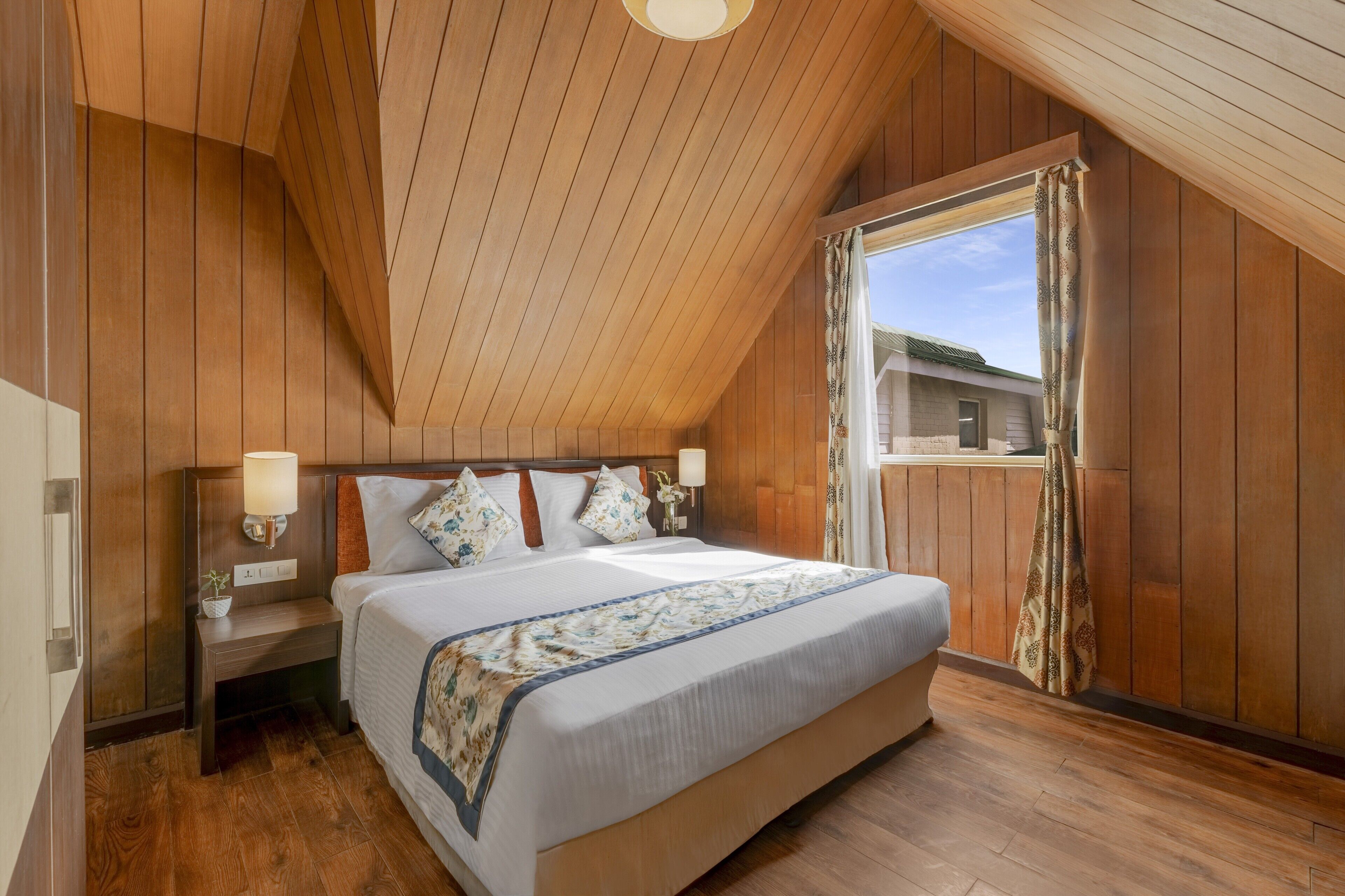 A bedroom with a wooden ceiling, a large bed with a patterned bedspread, two bedside tables with lamps, and a window with curtains.