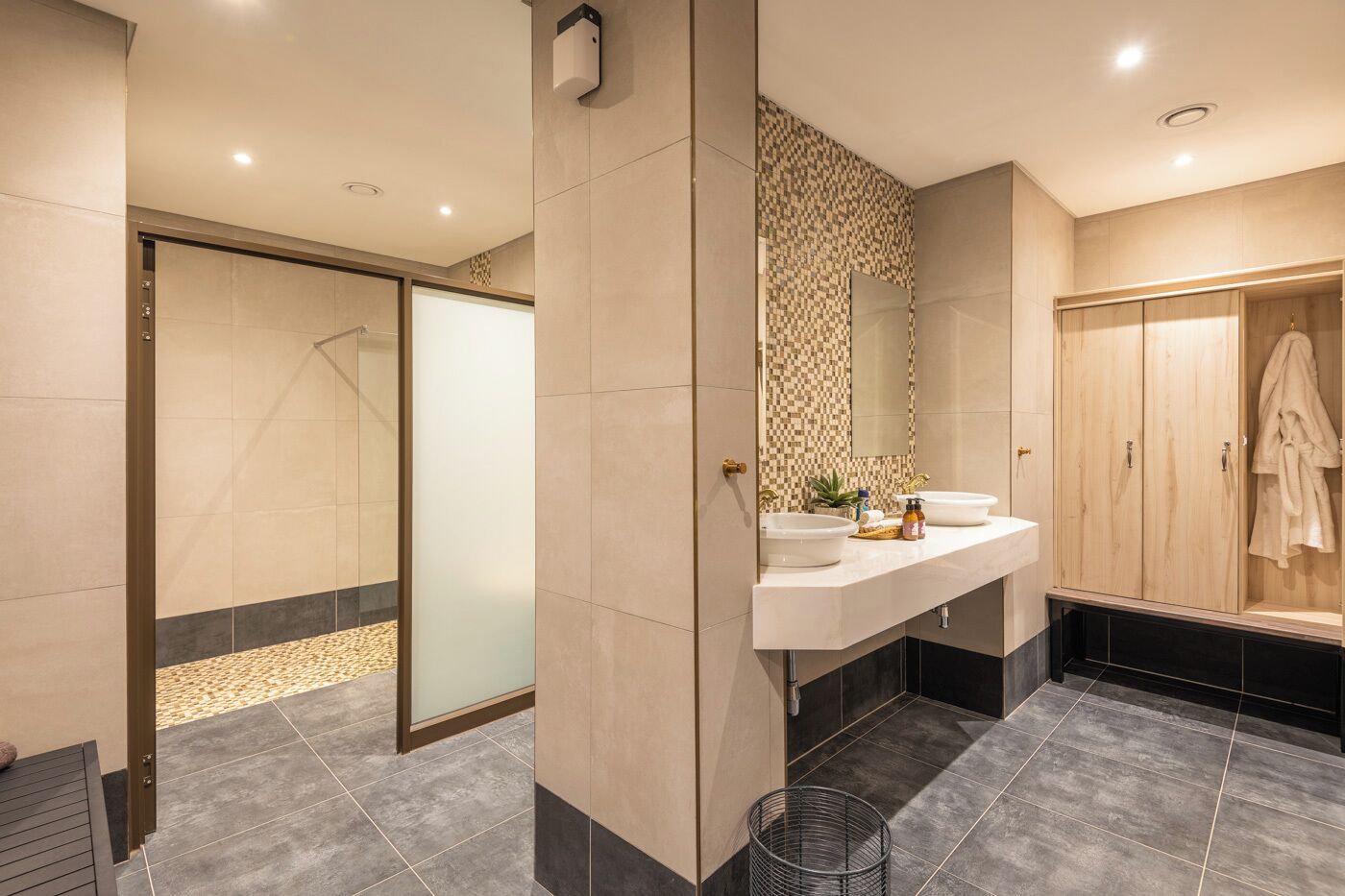 Une salle de bain moderne équipée d’une douche, de deux lavabos, d’un grand miroir et d’un porte-peignoir.
