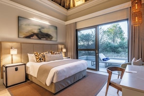A hotel room with a large bed, a desk, a chair, a suitcase, and a balcony with a view of trees. - Mokuti Etosha (Tsumeb)