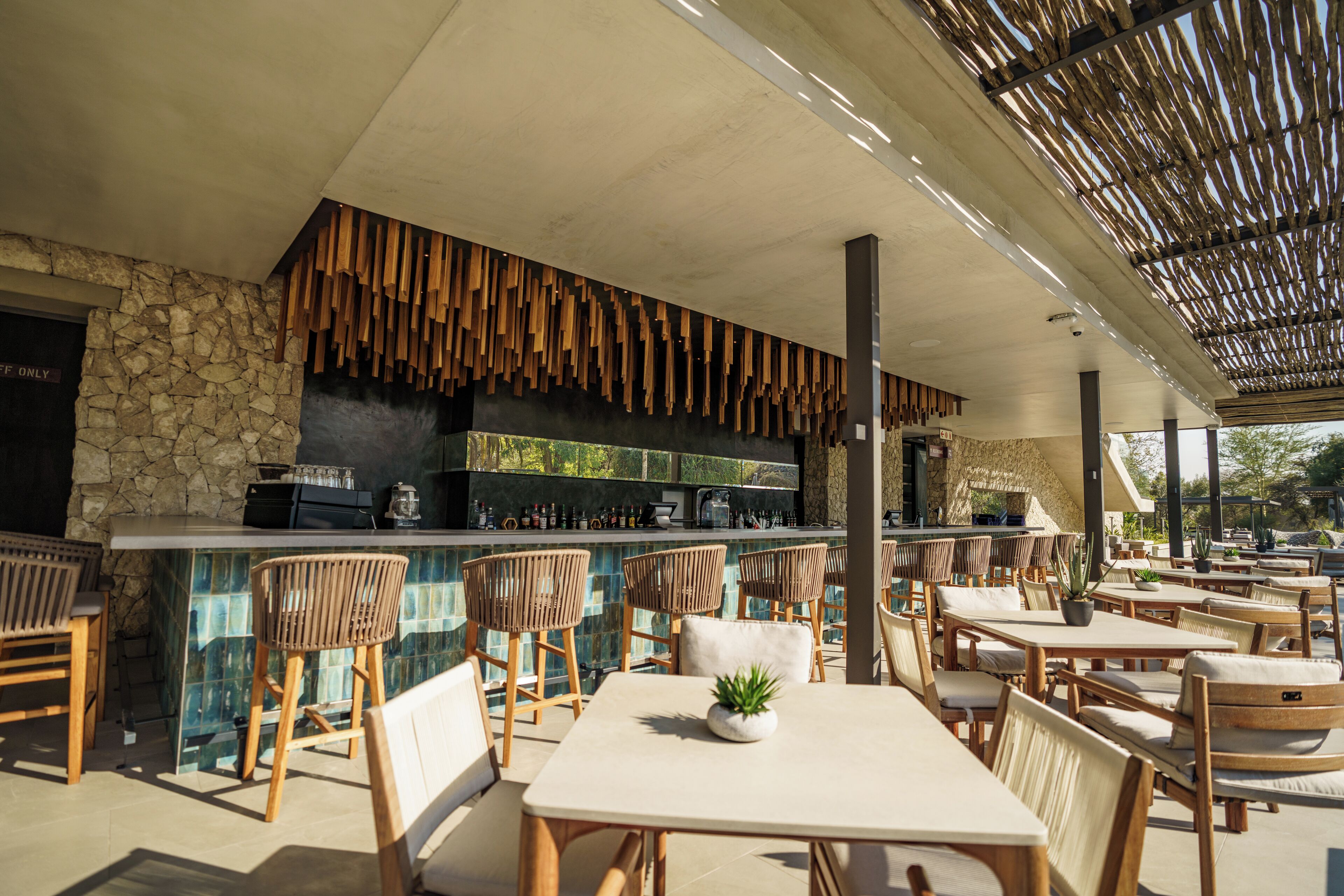 Un espacio de comedor al aire libre con mobiliario de madera, una barra con pared de piedra de fondo y techo de paja.