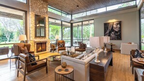 A spacious living area with a stone fireplace, comfortable seating, and large windows offering a view of the outdoors. - Mokuti Etosha (Tsumeb)