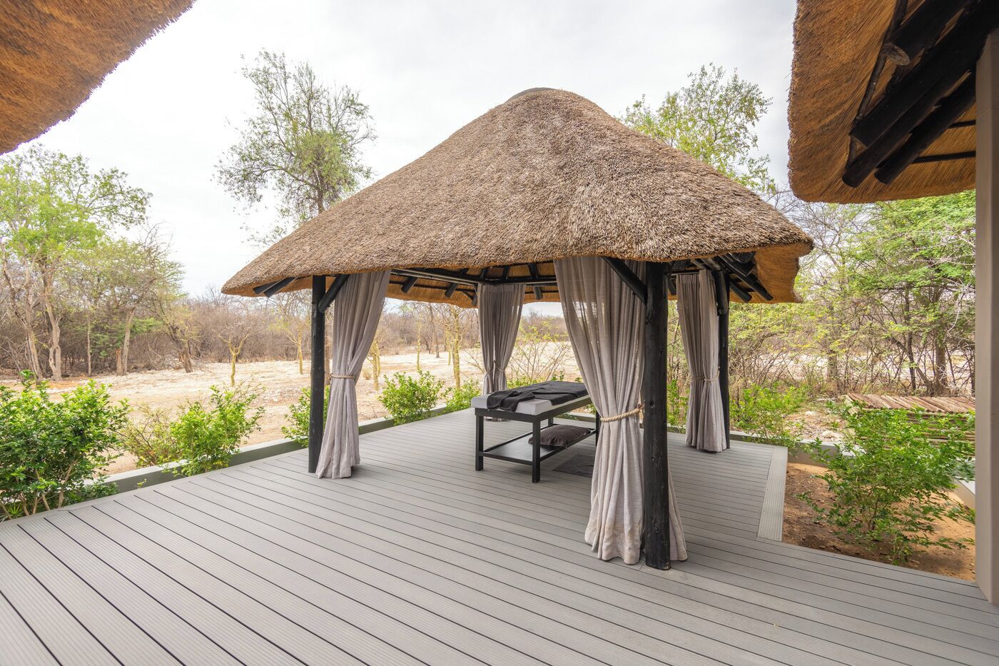 A thatched-roof gazebo with a bed and curtains, surrounded by a wooden deck and natural landscape.