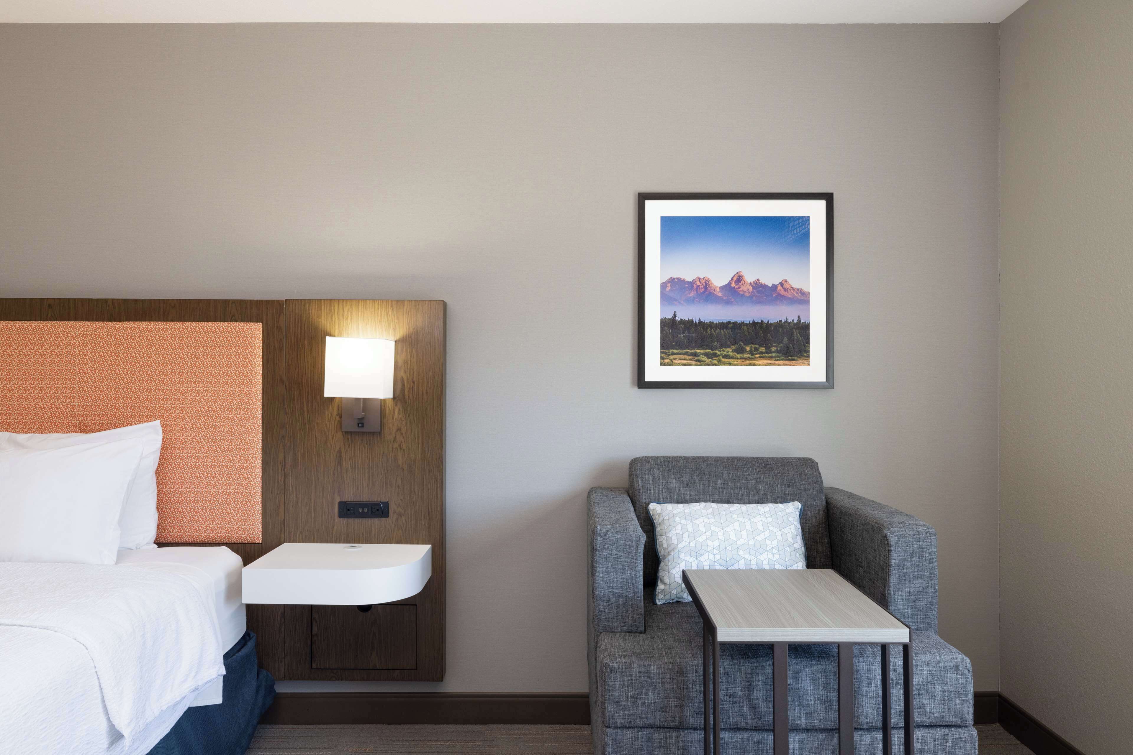 A hotel room with a bed, a nightstand with a lamp, a gray armchair, a small table, and a framed picture of mountains on the wall.