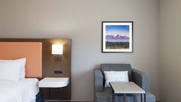 A hotel room with a bed, a nightstand with a lamp, a gray armchair, a small table, and a framed picture of mountains on the wall.