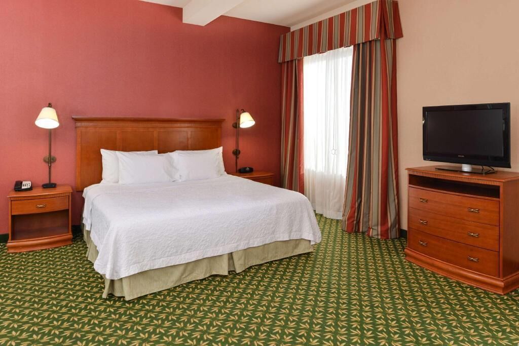 A hotel room with a bed, bedside lamps, a television, a dresser, and a window with curtains.