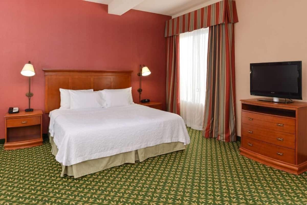 A hotel room with a bed, bedside lamps, a television, a dresser, and a window with curtains.