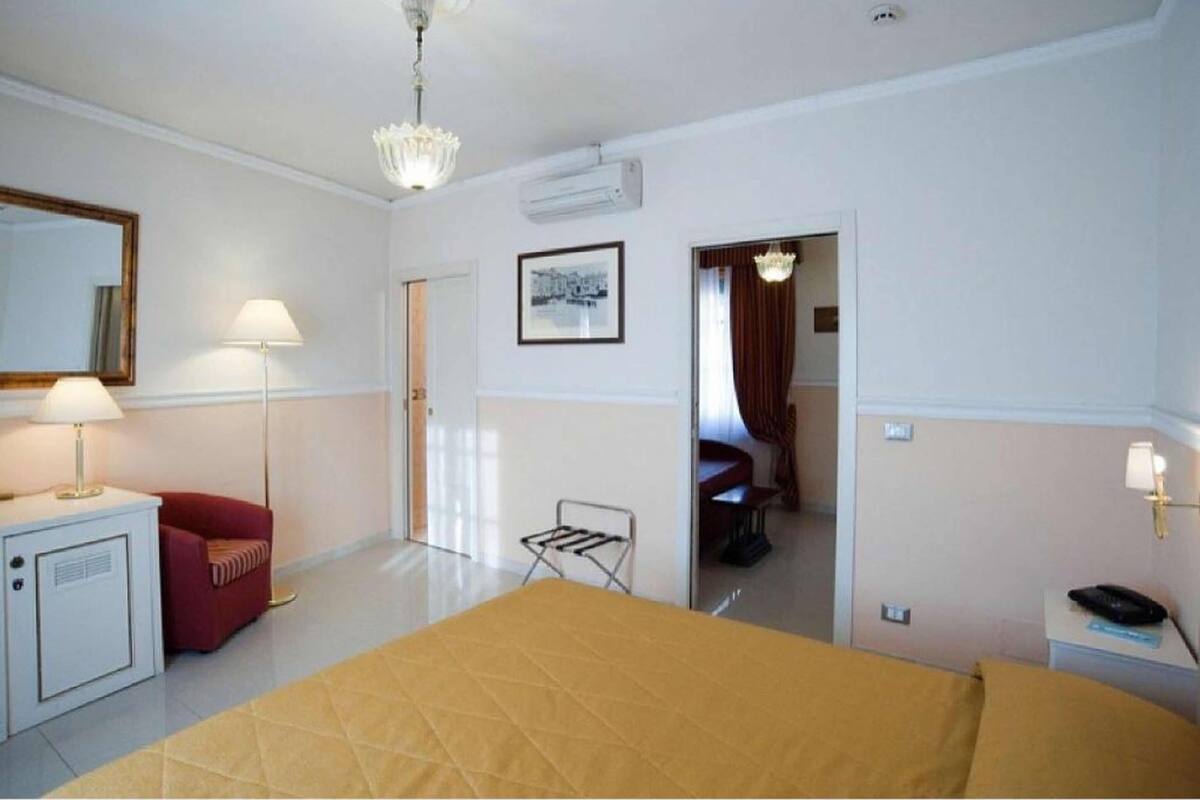Une chambre d’hôtel avec un lit, un bureau avec une lampe, une chaise rouge, une petite table avec une lampe, un tableau au mur et un appareil de climatisation.