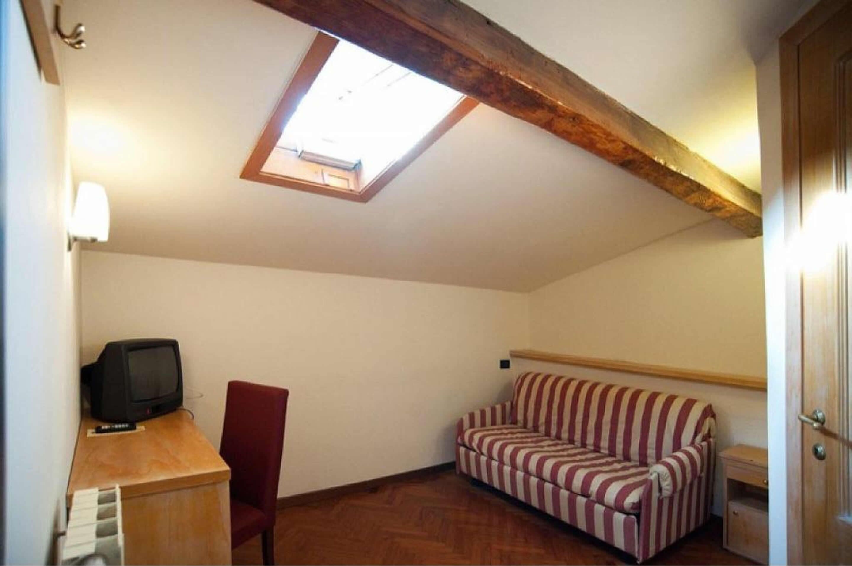 Une petite pièce chaleureuse avec un plafond en pente, un seul fauteuil rouge, un canapé à rayures, un téléviseur posé sur une table en bois et une lucarne.