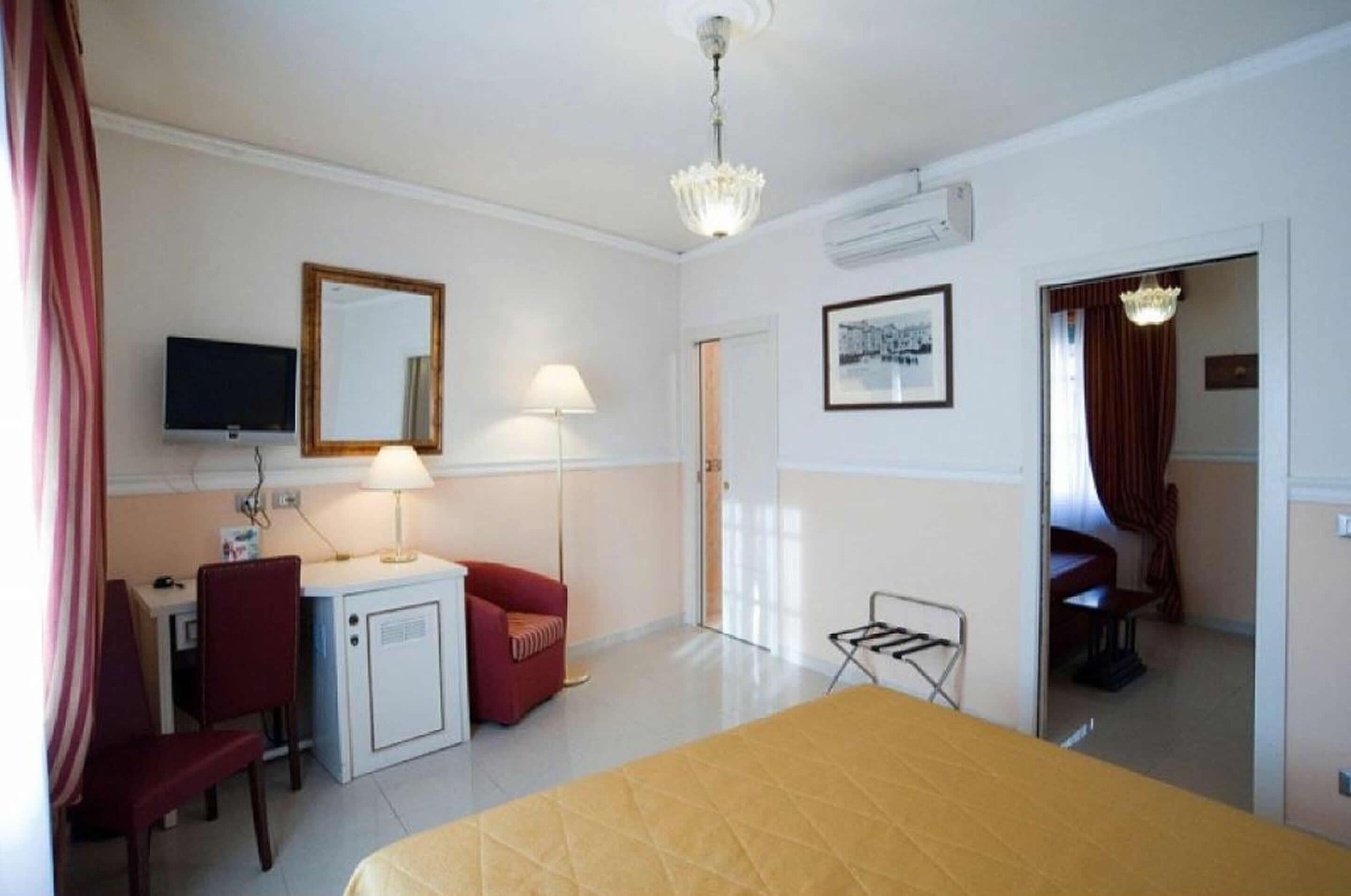 Una camera d'albergo con un letto, una scrivania con una sedia, una televisione, una lampada, uno specchio e una porta che dà su un'altra stanza.