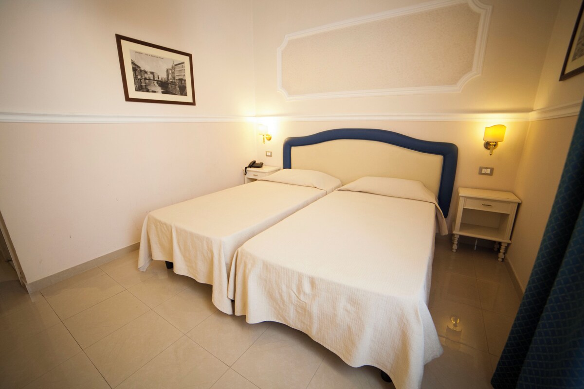 Ein Hotelzimmer mit zwei Einzelbetten, einem Nachttisch mit Lampe, einem gerahmten Bild an der Wand und einem gefliesten Boden.