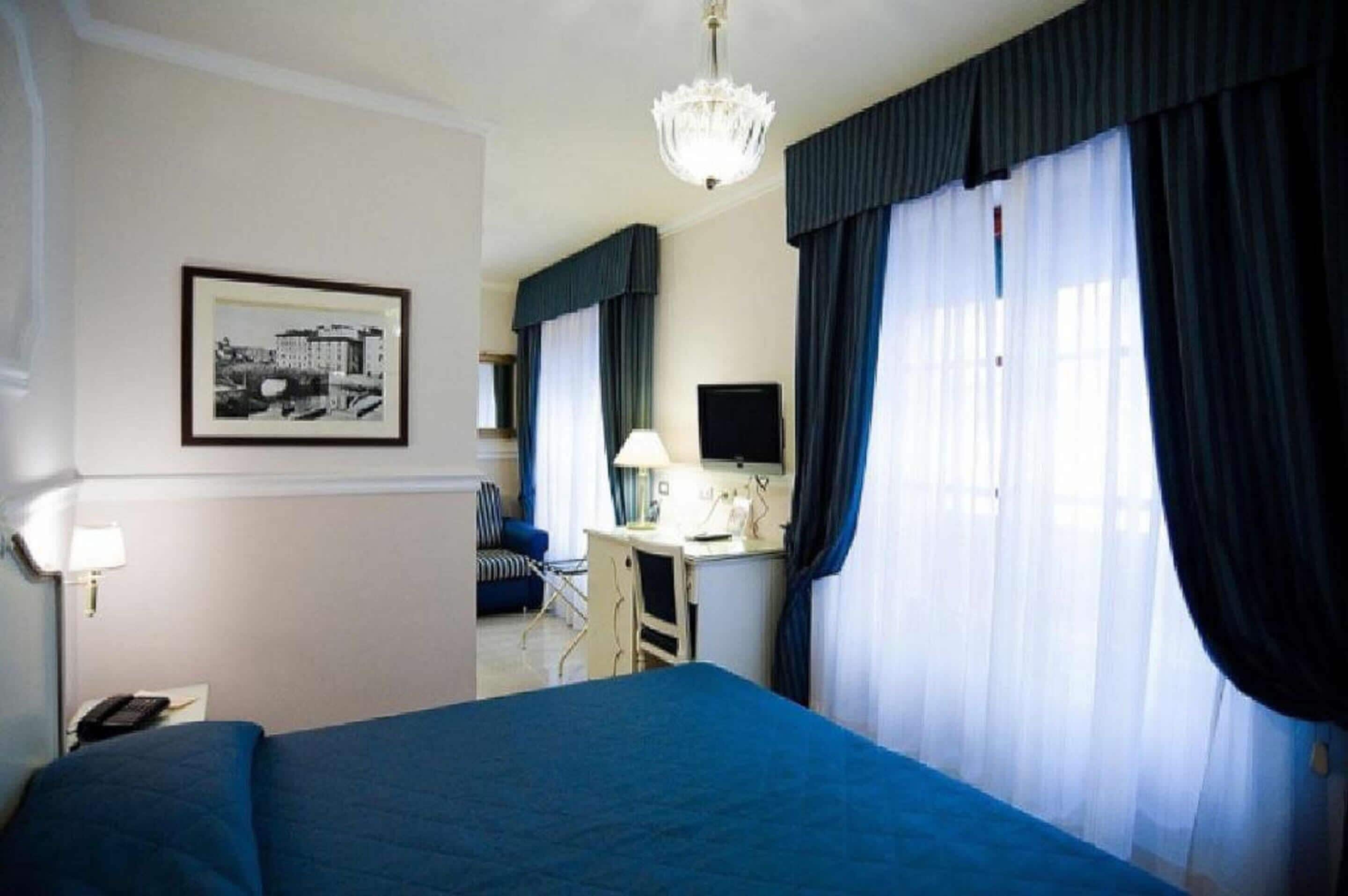 A hotel room with a bed, a desk, a chair, a television, and a chandelier.