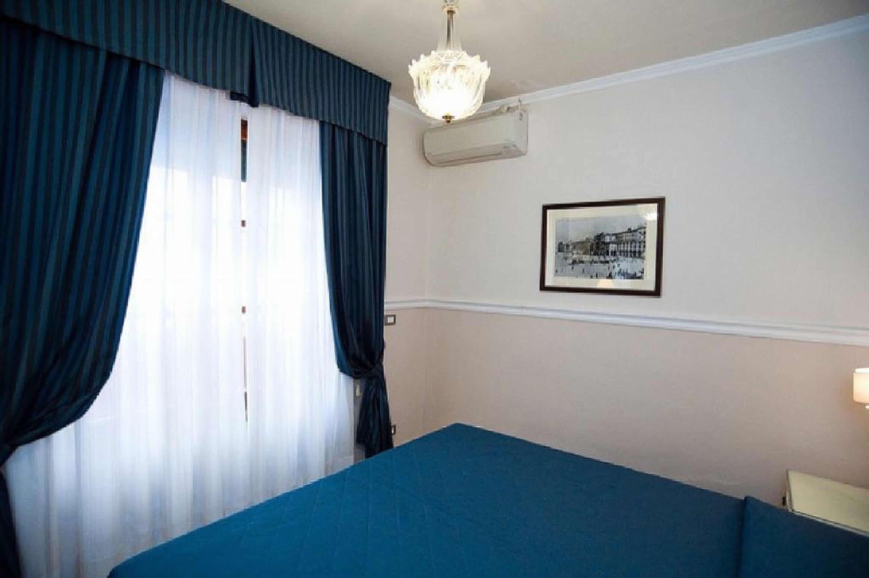 A hotel room with a bed, a window with curtains, a wall-mounted air conditioner, a framed picture, and a chandelier.