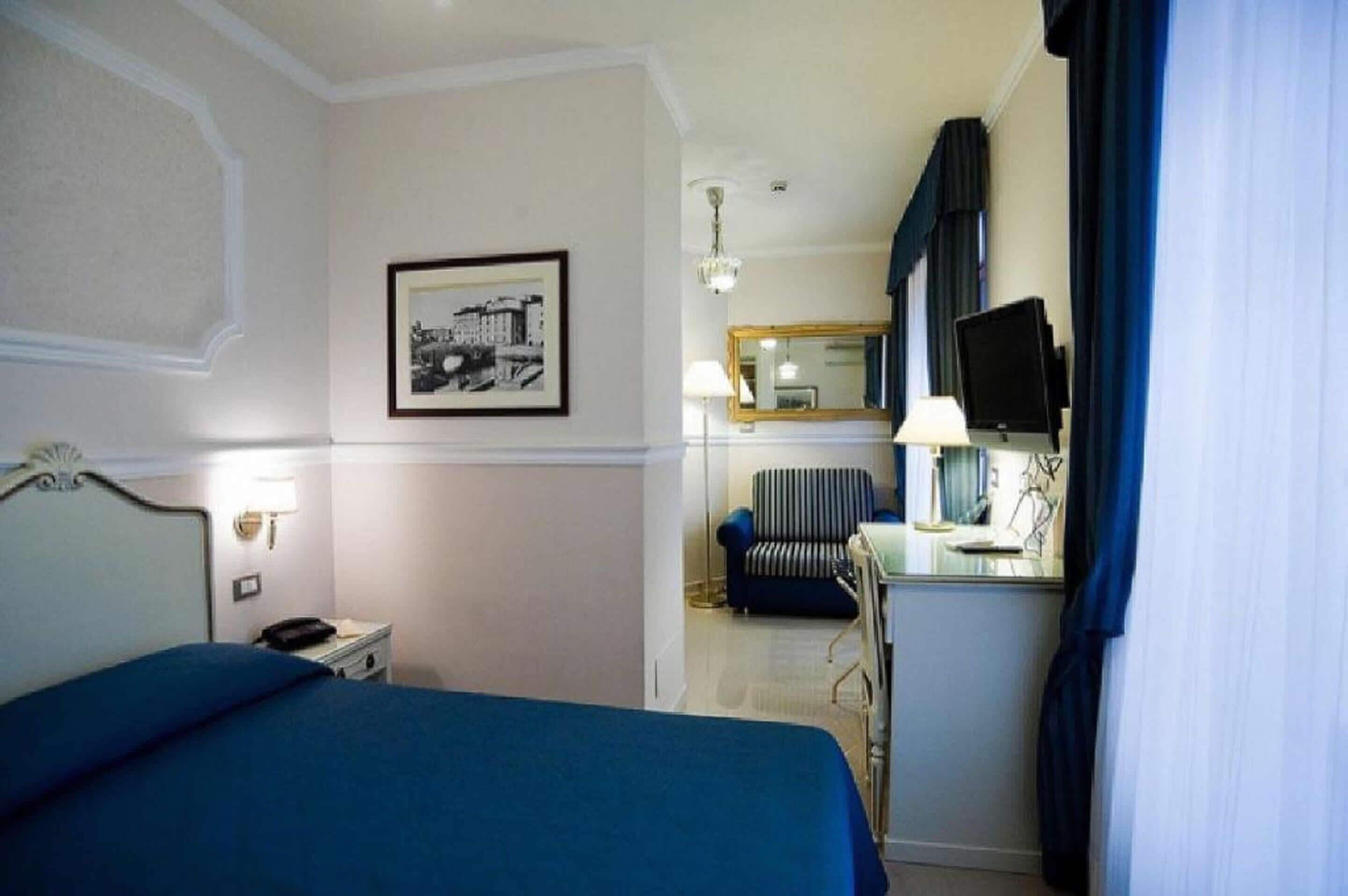 A hotel room with a bed, a desk with a lamp, a chair, a television, and a framed picture on the wall.