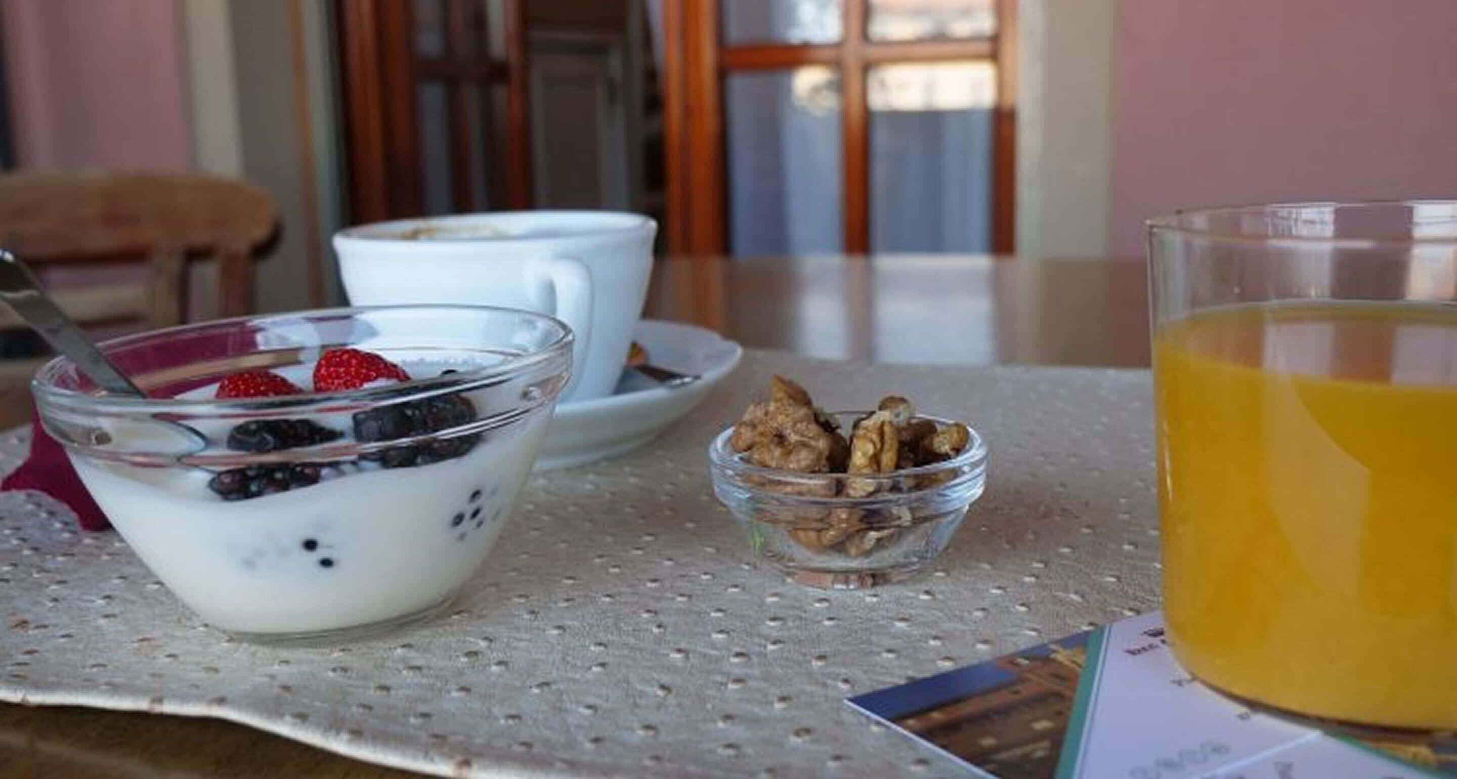 Uma mesa com um recipiente de vidro com iogurte coberto de frutas vermelhas, um pequeno pote de granola, uma xícara de café e um copo de suco de laranja.