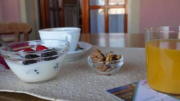 Uma mesa com um recipiente de vidro com iogurte coberto de frutas vermelhas, um pequeno pote de granola, uma xícara de café e um copo de suco de laranja.