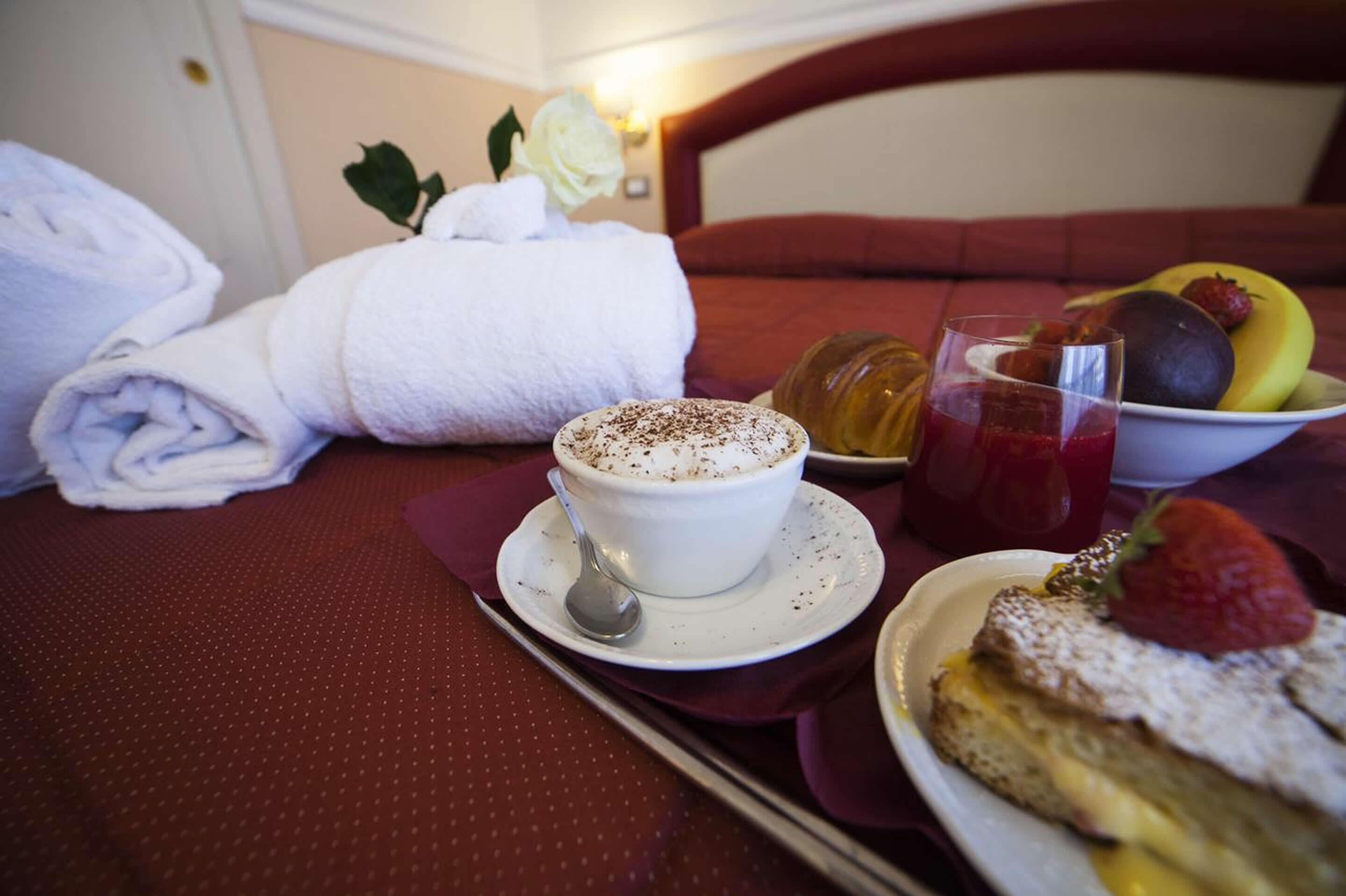 A neatly made bed with white towels, a cup of coffee with whipped cream, a plate of fruit, and a glass of juice on a table.