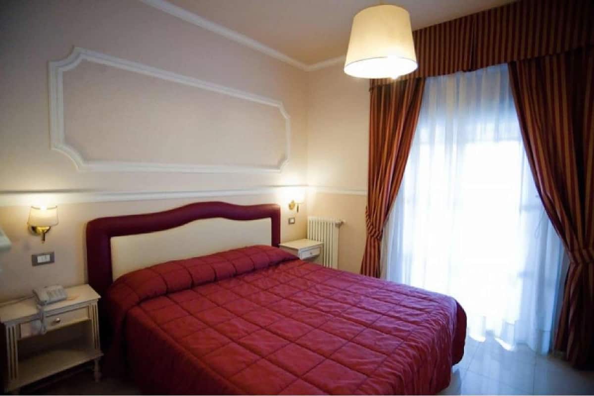 Une chambre d’hôtel avec un grand lit, une table de chevet avec une lampe, un radiateur et une fenêtre avec des rideaux.
