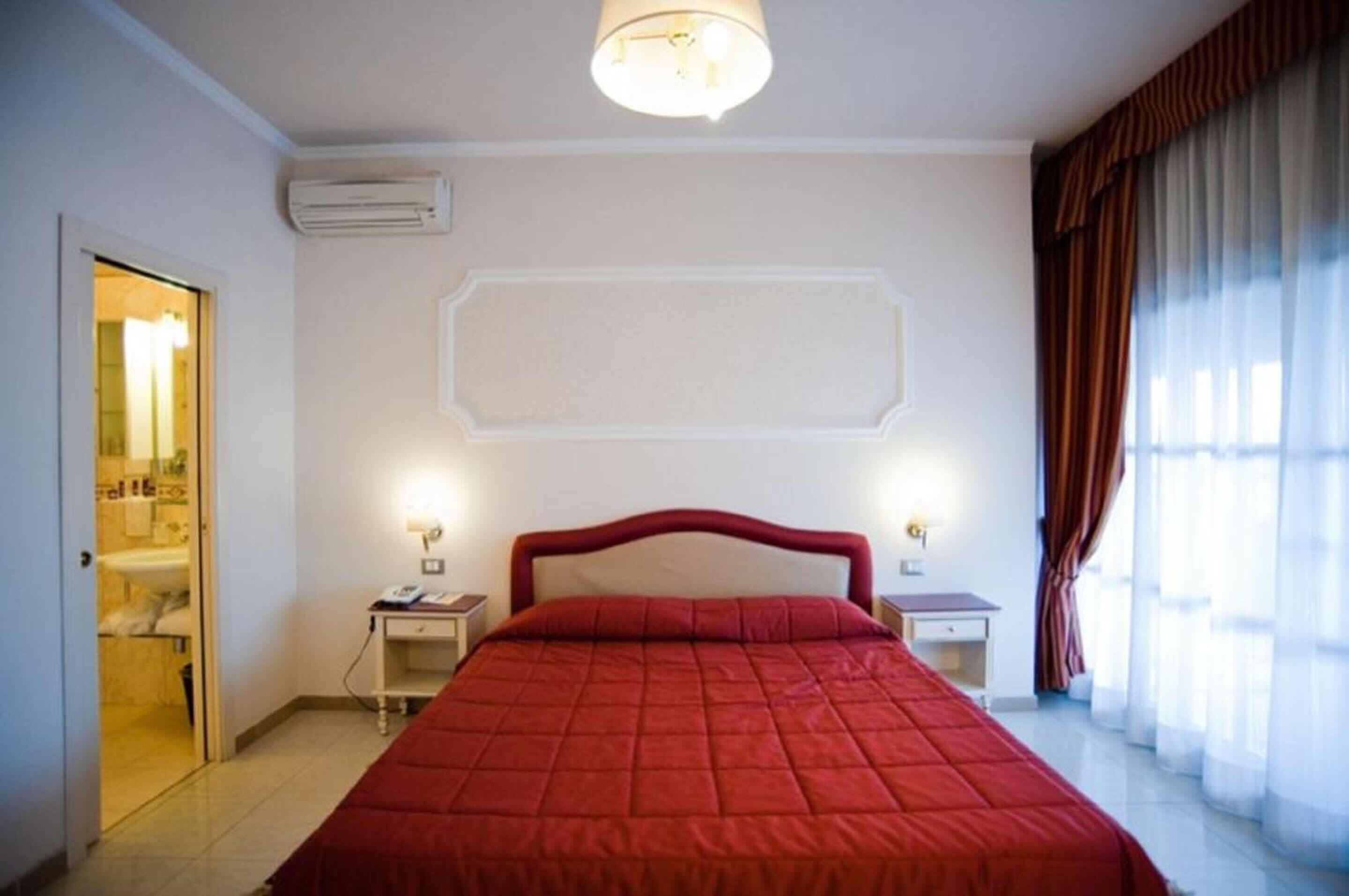 A hotel room with a large bed, a bedside table, a nightstand, an air conditioning unit, and a bathroom visible through an open door.