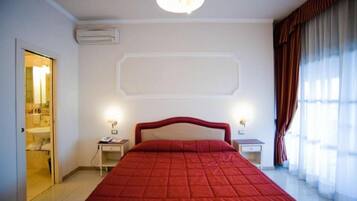 A hotel room with a large bed, a bedside table, a nightstand, an air conditioning unit, and a bathroom visible through an open door.