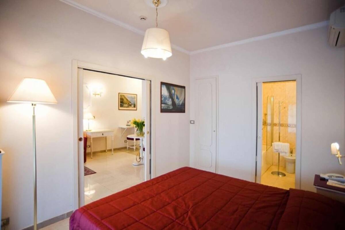 Une chambre d’hôtel avec un lit, un bureau, une chaise, une lampe et une salle de bain visible à travers une porte ouverte.