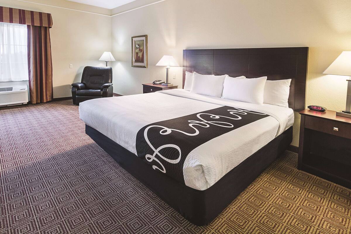 Un dormitorio de hotel con una cama grande, una silla, dos mesitas de noche con lámparas, un cuadro en la pared y una ventana con cortinas.