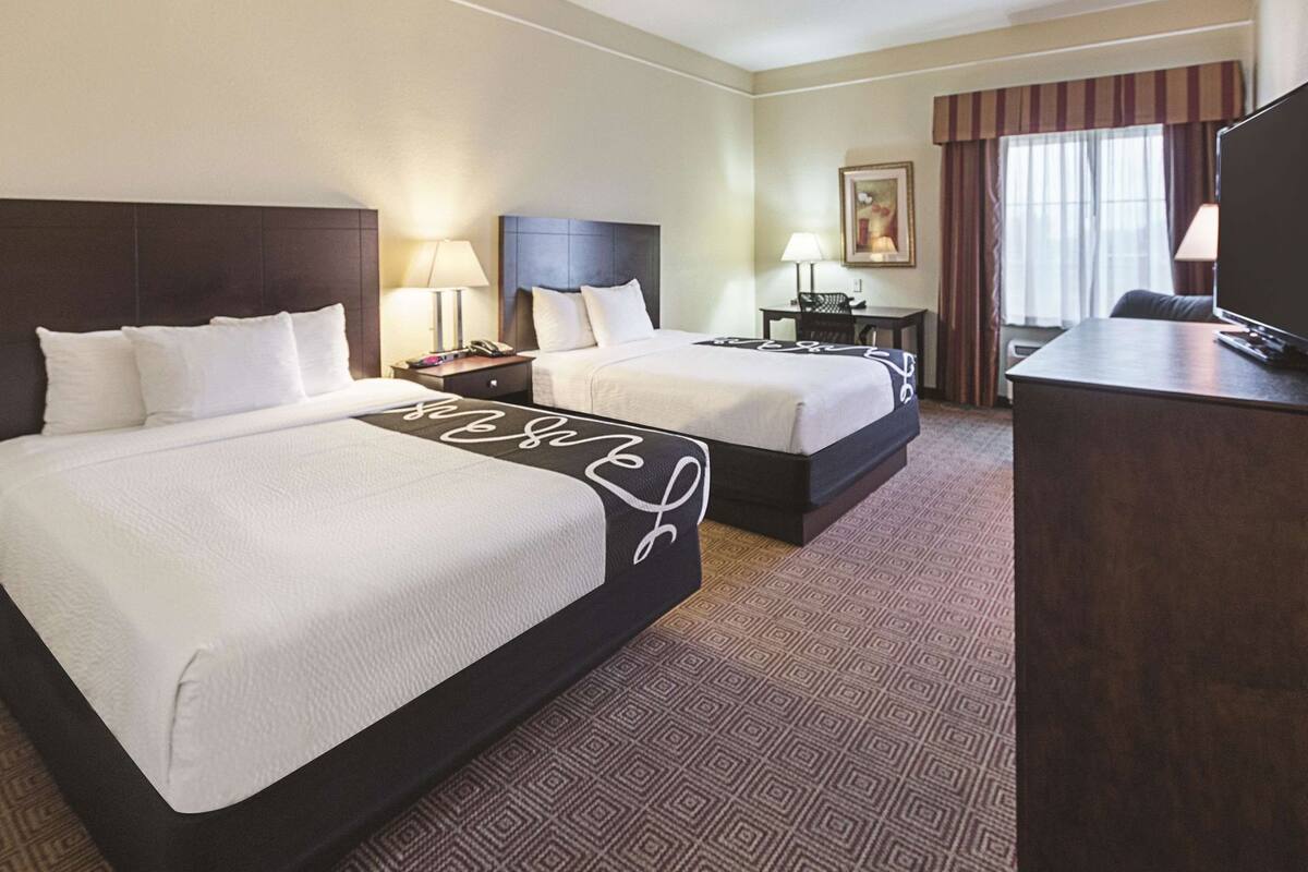 Habitación de hotel con dos camas, un escritorio, una silla, una lámpara y un cuadro en la pared.