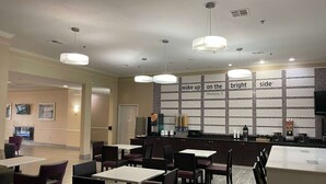 Interior modern d'un restaurant amb diverses taules i caires, una barra amb un quadre de menú, i il·luminació penjant.