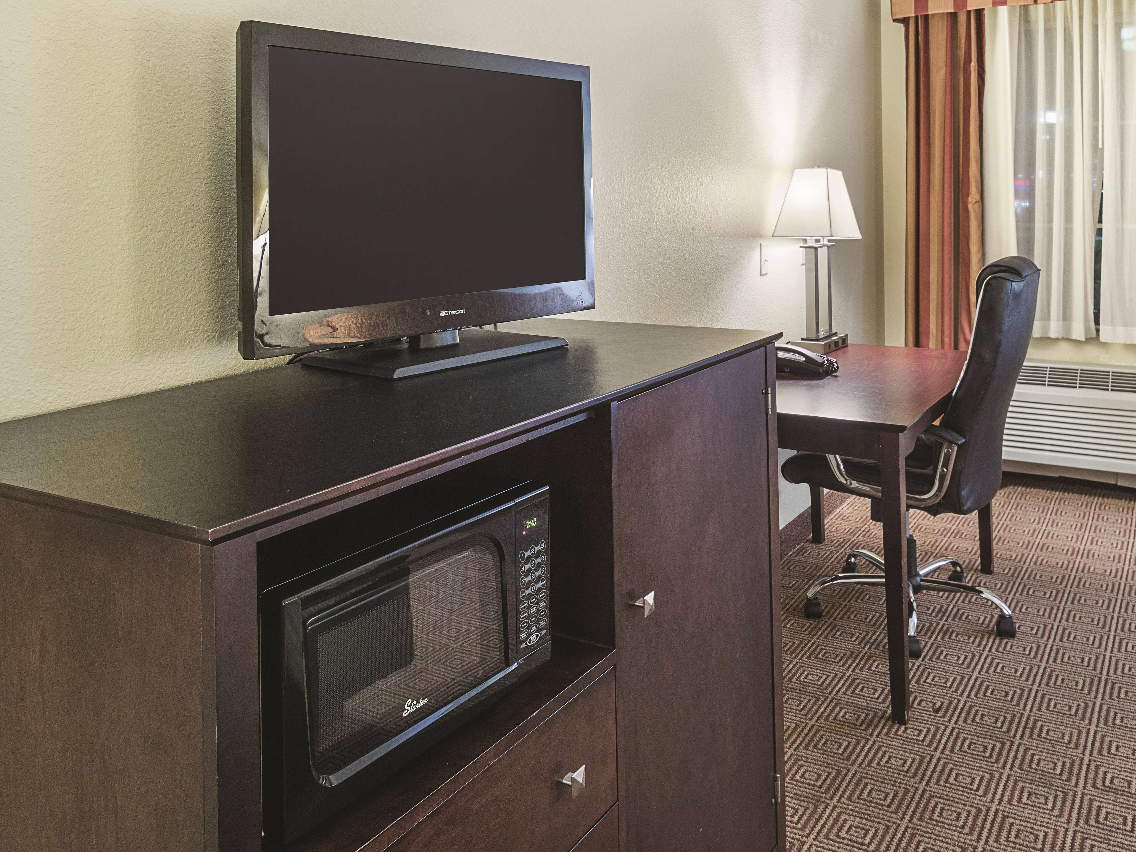 A hotel room with a flat-screen TV, a microwave, a desk, a chair, and a lamp.