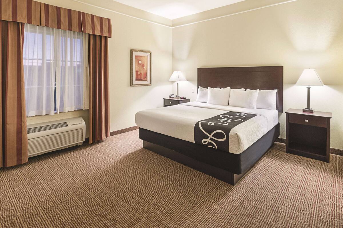 Habitación de hotel con una cama grande, dos mesitas de noche con lámparas, una ventana con cortinas, un cuadro en la pared y suelo alfombrado.