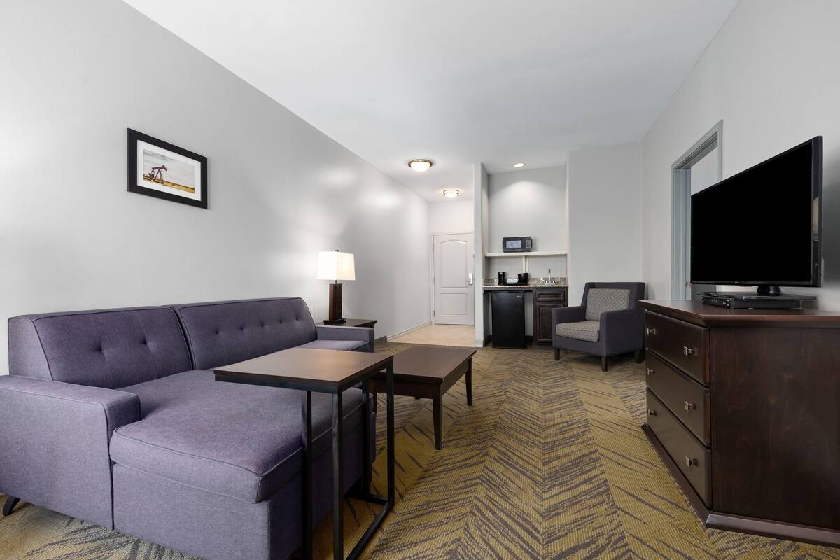 A modern hotel room with a sectional sofa, a flat-screen TV, a dining table, a microwave, and a kitchenette.